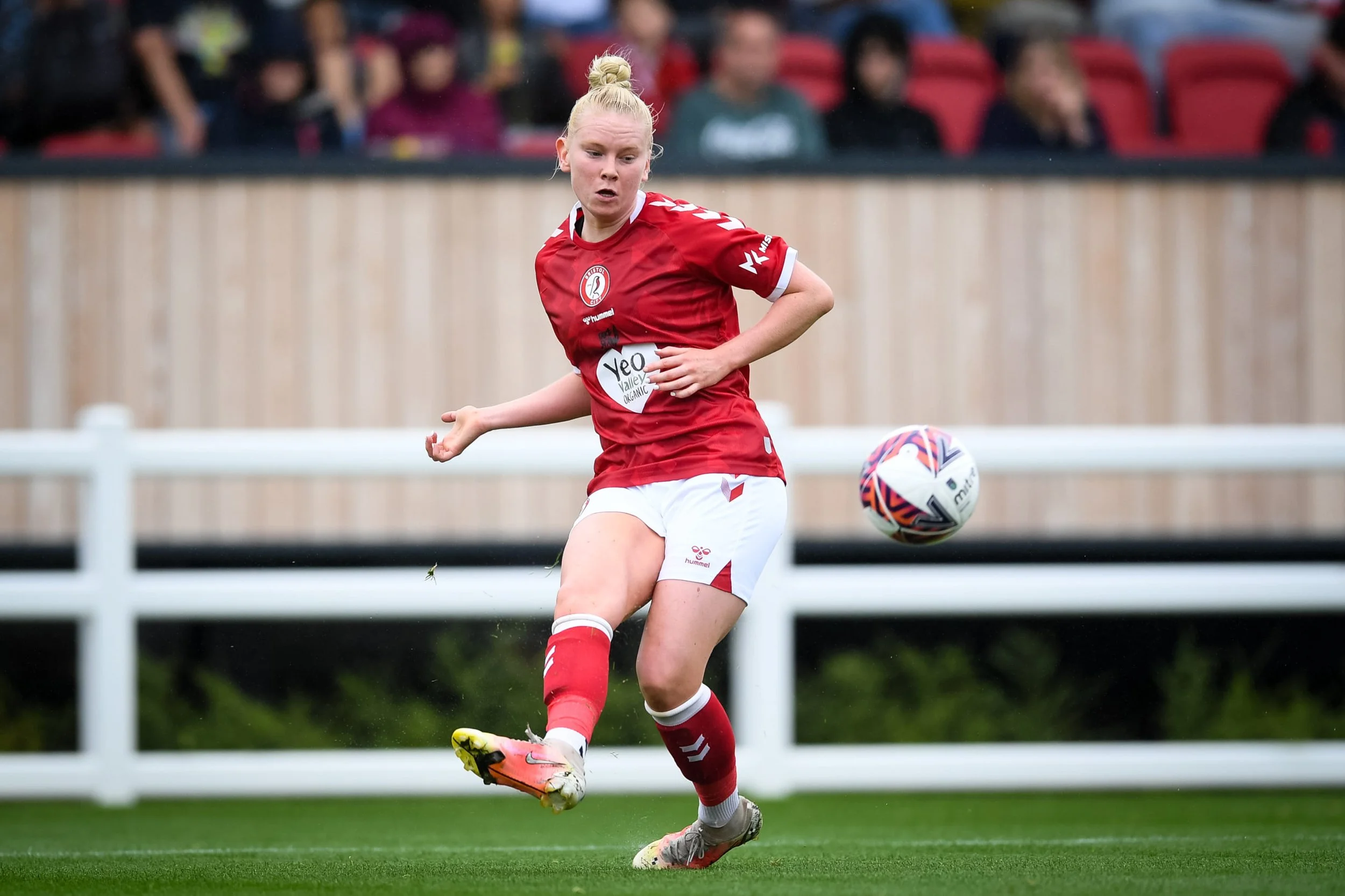 A female soccer player in a red jersey and white shorts is kicking a soccer ball on the field during a game. She has blond hair tied in a bun and is focused on the ball. Spectators are visible in the background.