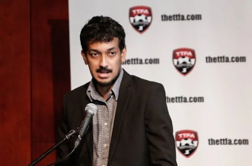A man with dark hair and a mustache standing at a microphone in front of a backdrop with the logo and website 'thetta.com' repeated.
