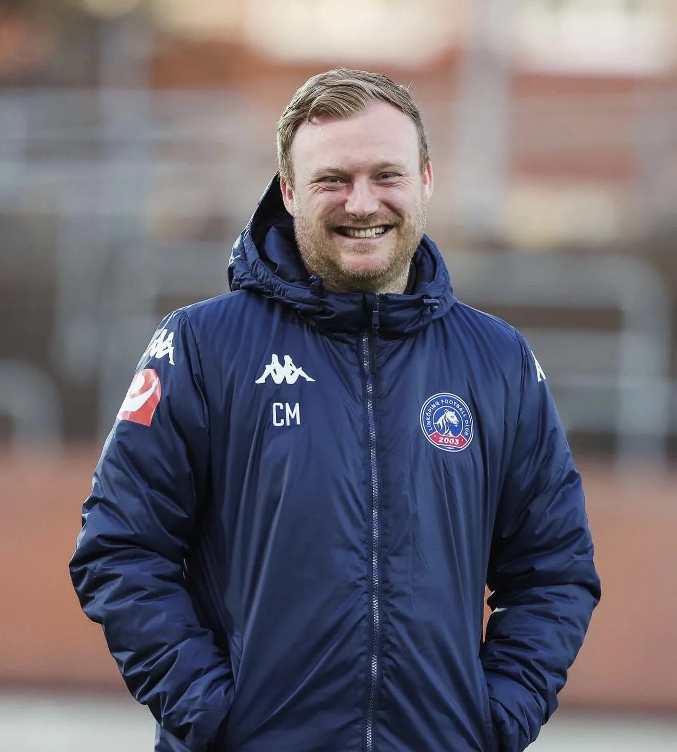 A man smiling, wearing a blue sports jacket with football club logos, with hands in pockets, standing outdoors with blurred background.