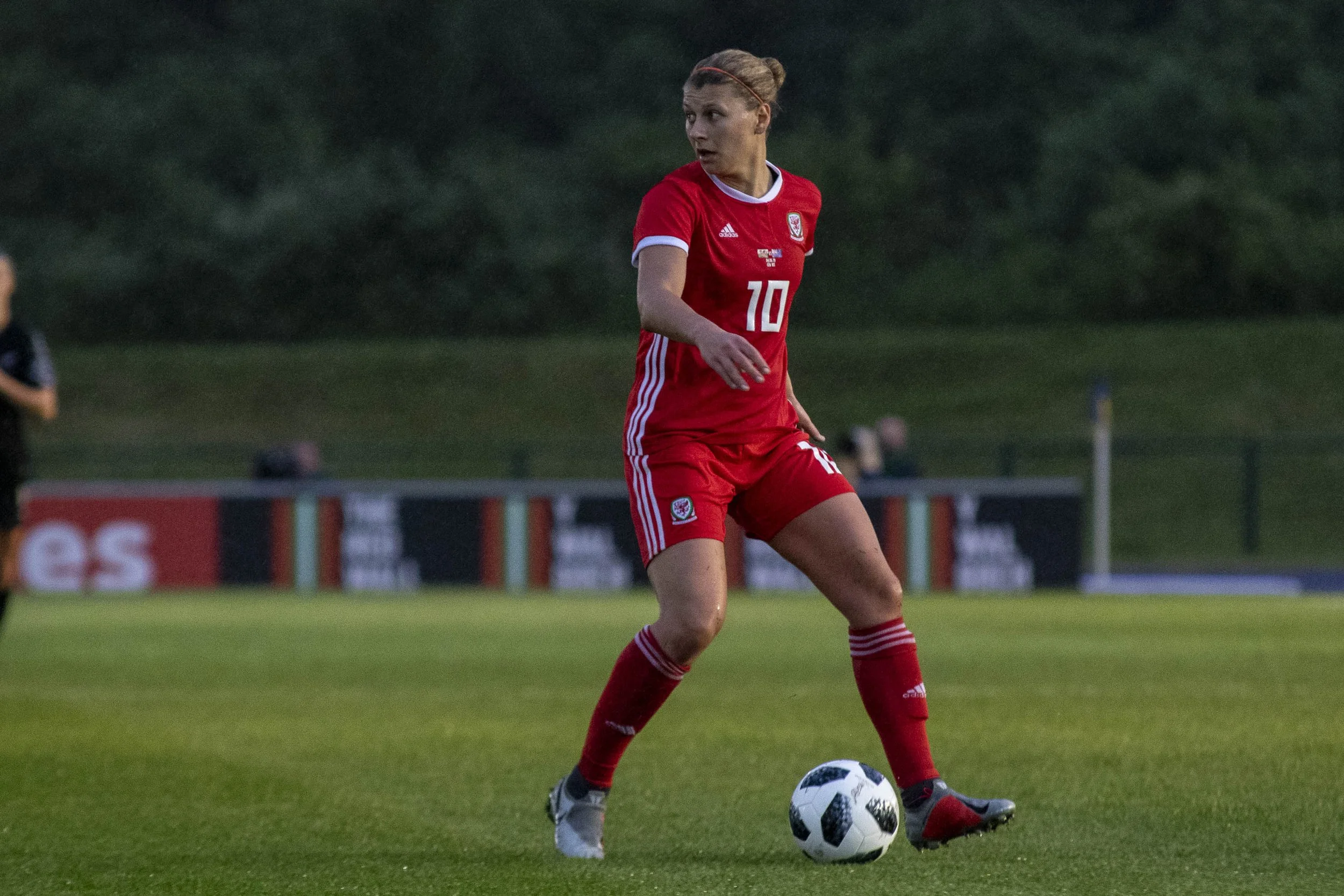 A female soccer player with a headband, wearing a red jersey with the number 10 and white accents, is standing on a grassy field, preparing to kick a black and white soccer ball.