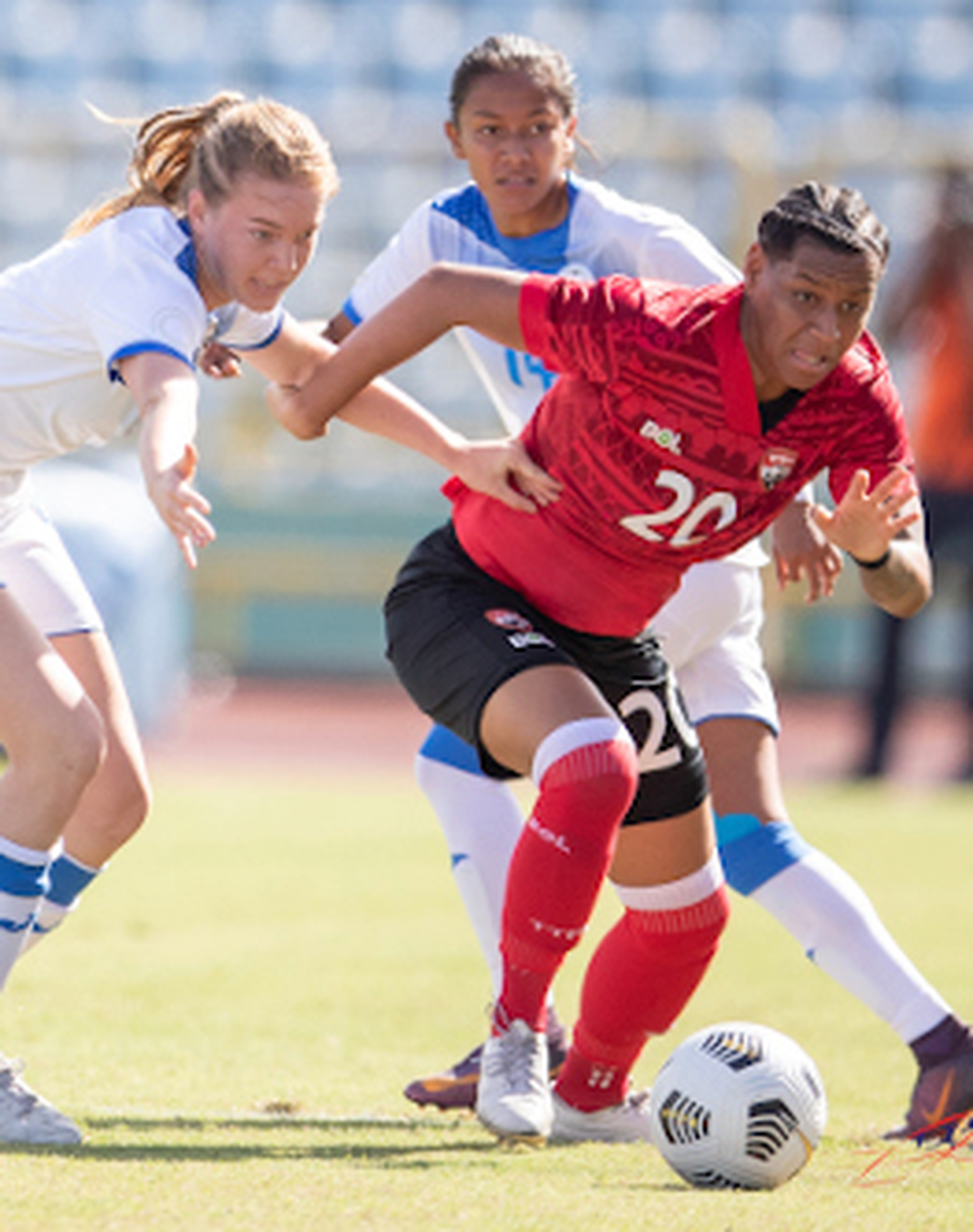 Three female soccer players competing for ball on field, two in white and one in red jersey, during match.