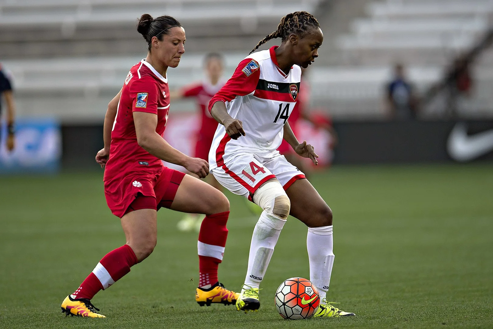 Two female soccer players, one in a red uniform and one in a white uniform, compete for the ball on the field. The player in white is wearing a knee brace.