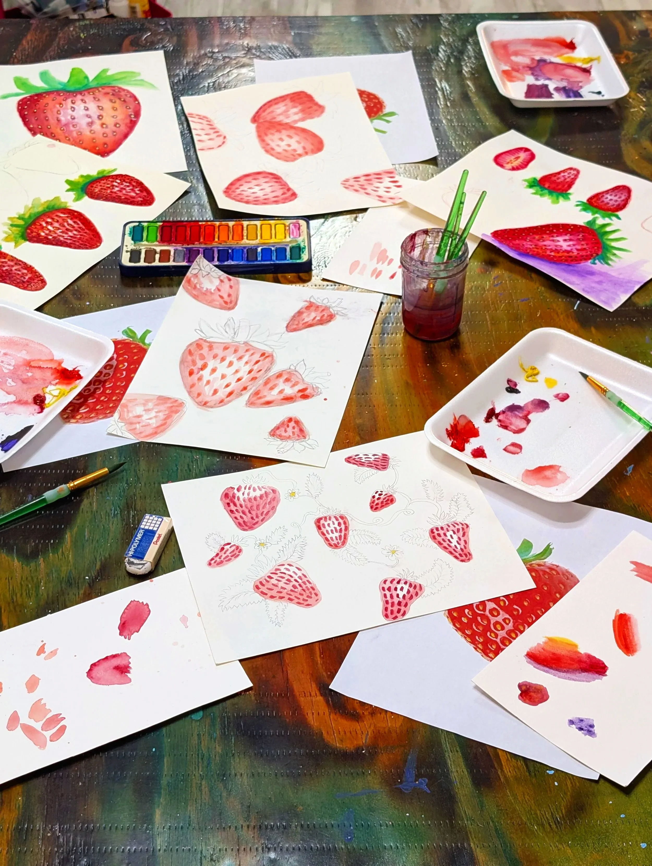 Watercolor painting supplies and strawberry illustrations on paper, including painted strawberries, a paint palette, and brushes, on a colorful wooden table.