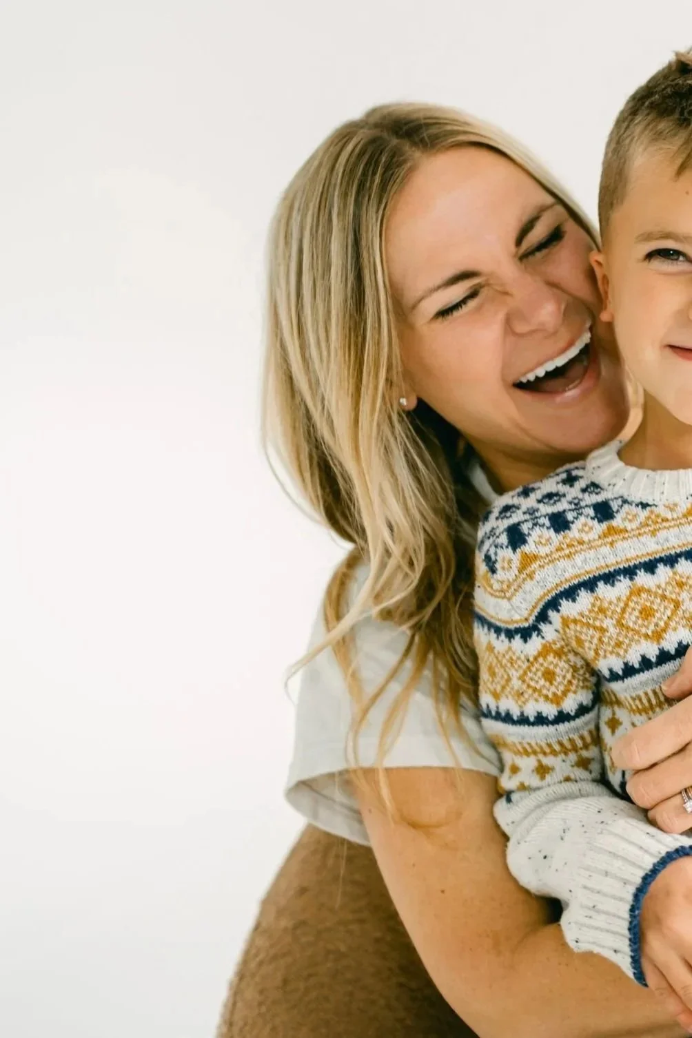 A woman with long blonde hair embracing a young boy, both smiling and laughing against a plain white background.