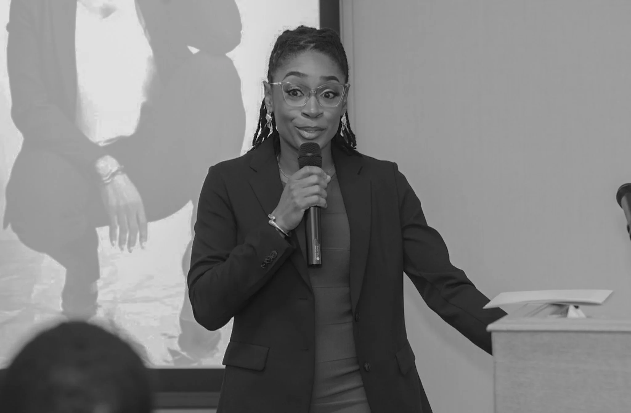 A woman with glasses speaking into a microphone at an indoor event with a presentation slide projected behind her.