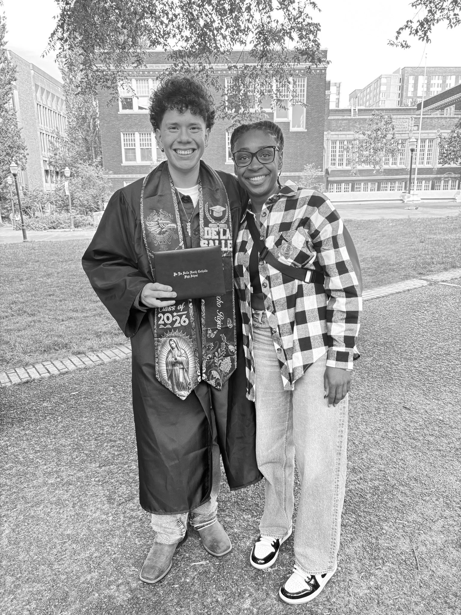 Two women standing outdoors on grass, smiling for the camera, with buildings and trees in the background.