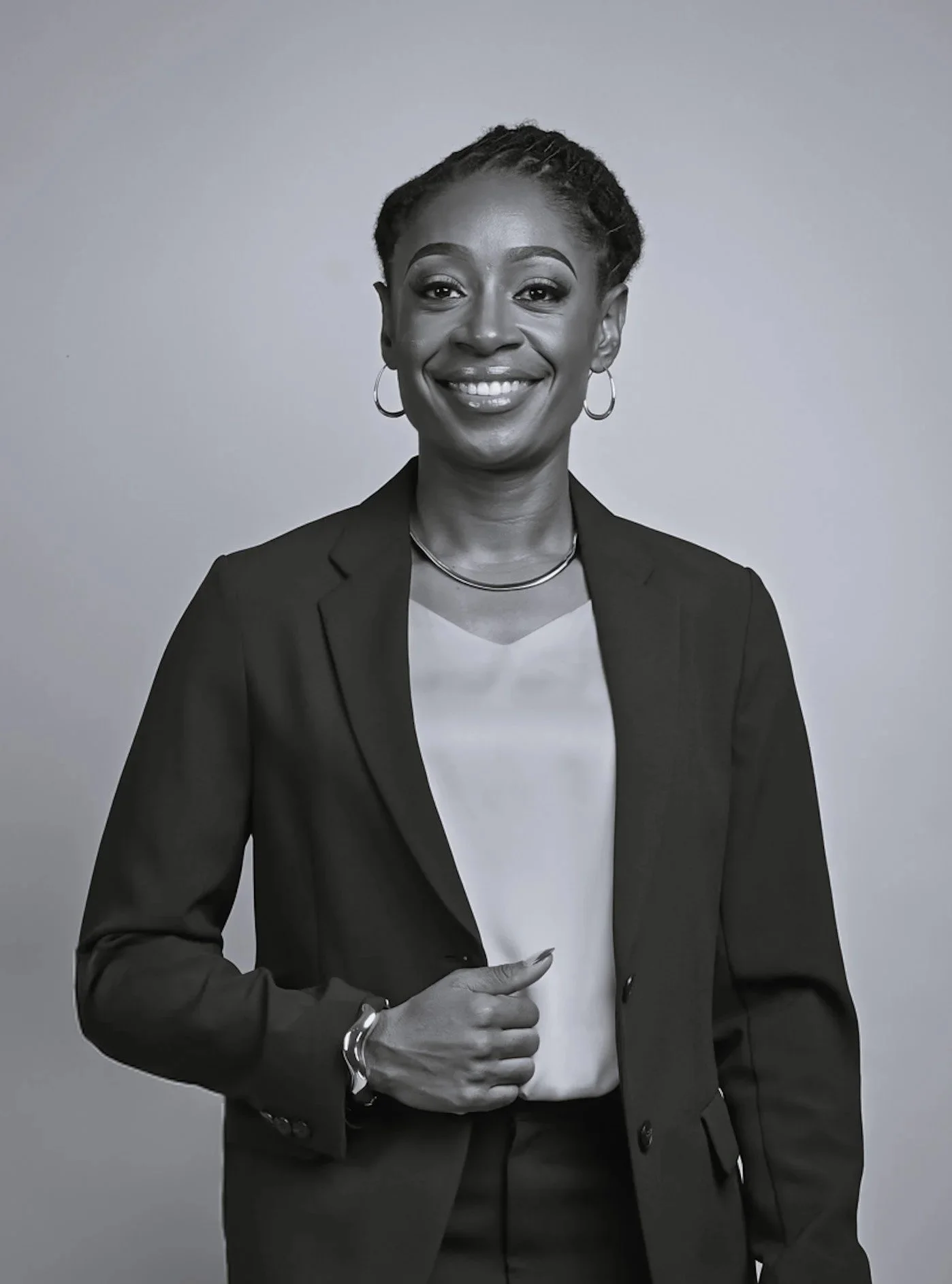 Black and white portrait of a woman smiling, wearing a blazer, white blouse, hoop earrings, and a necklace, with her hand resting on her waist.