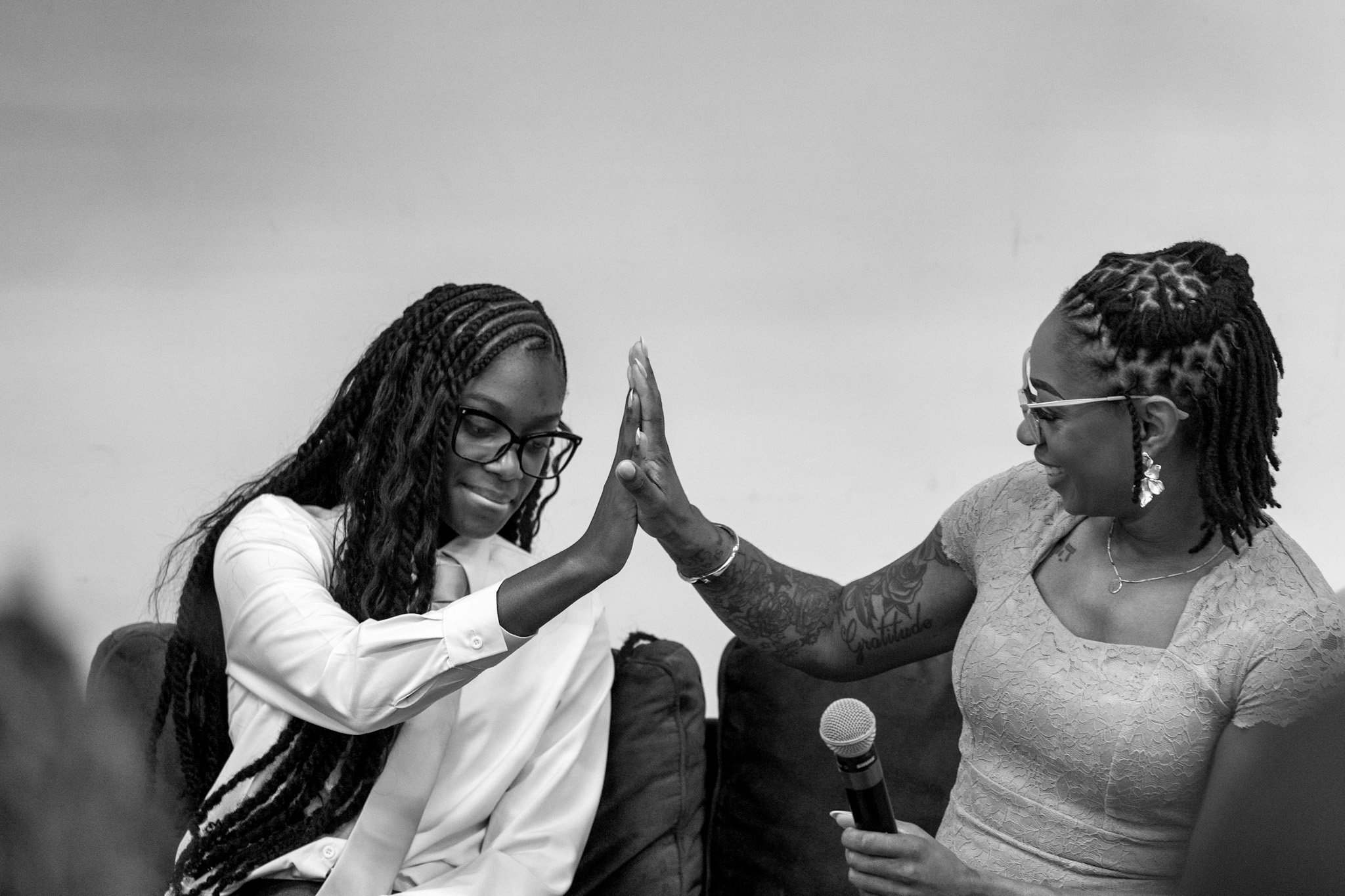 Two women with braided hair and glasses exchange a high-five, smiling at each other, with one holding a microphone. The woman on the right has tattoos on her arms and wears earrings, while the woman on the left has a white shirt.