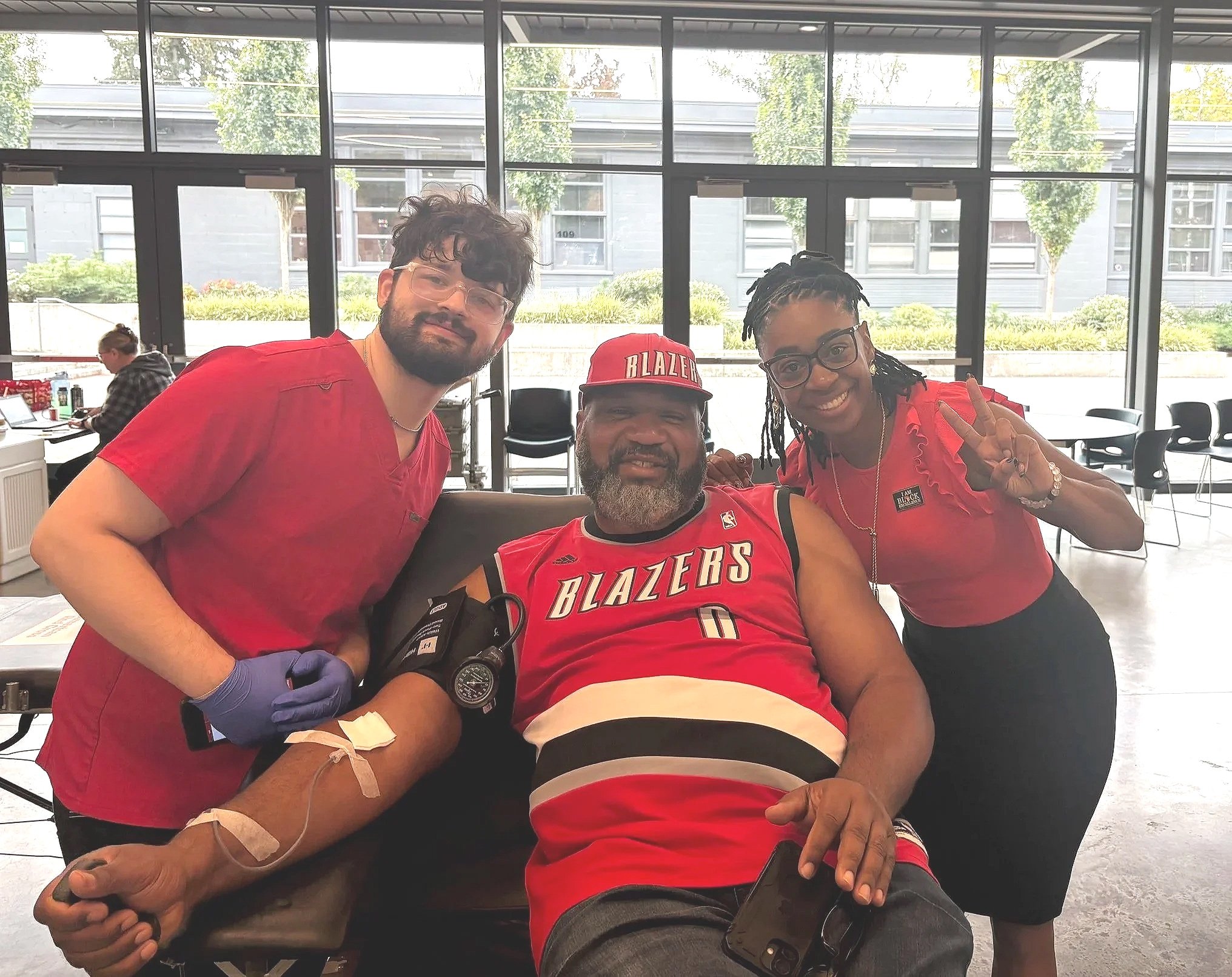 Three people, including a man in a Portland Trail Blazers basketball jersey, sitting at a table during a blood donation. The man is smiling with a blood pressure cuff on his arm. Two women, one making a peace sign, are standing beside him in an indoor space with large windows showing trees outside.