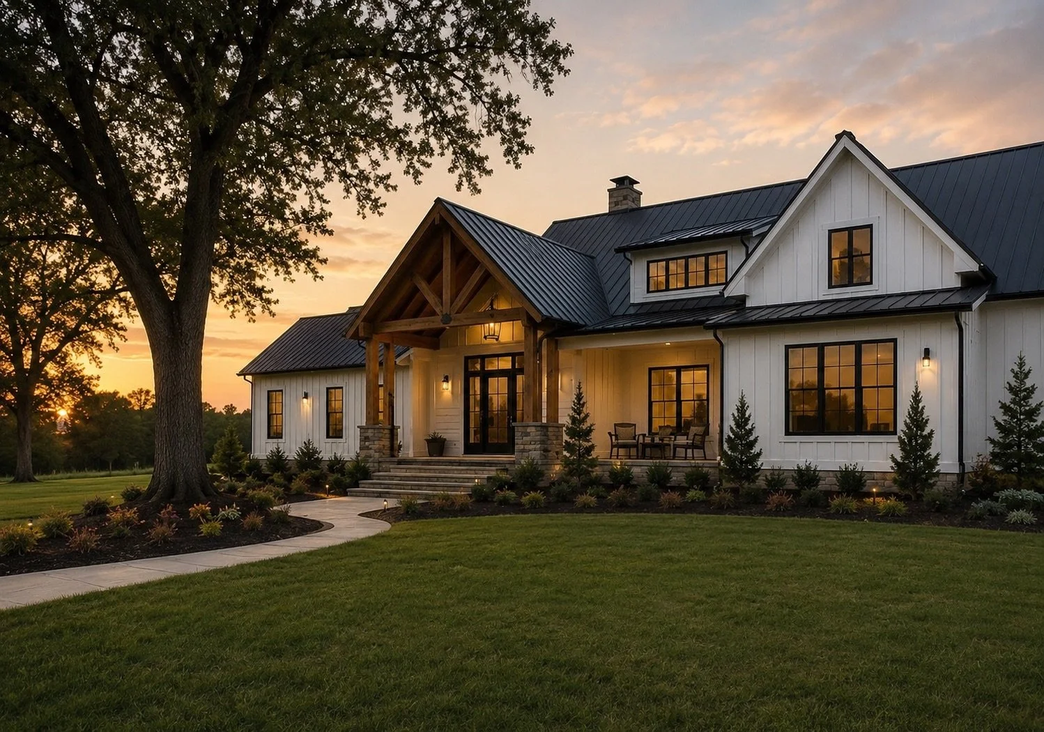 A modern farmhouse style home with white siding, black window frames, and a dark metal roof, nestled in a well-manicured yard with a large tree and sunset in the background.