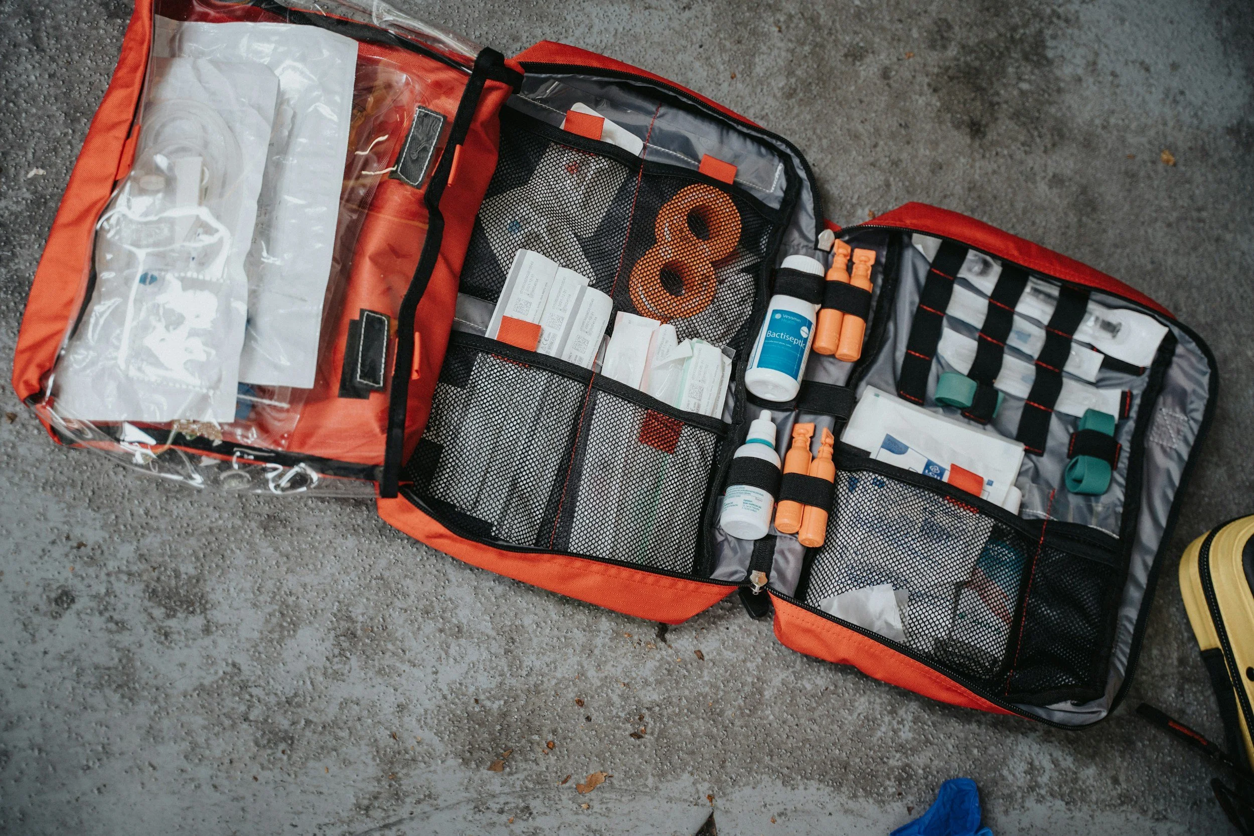 Open emergency medical kit bag on gravel ground containing medical supplies and medications.