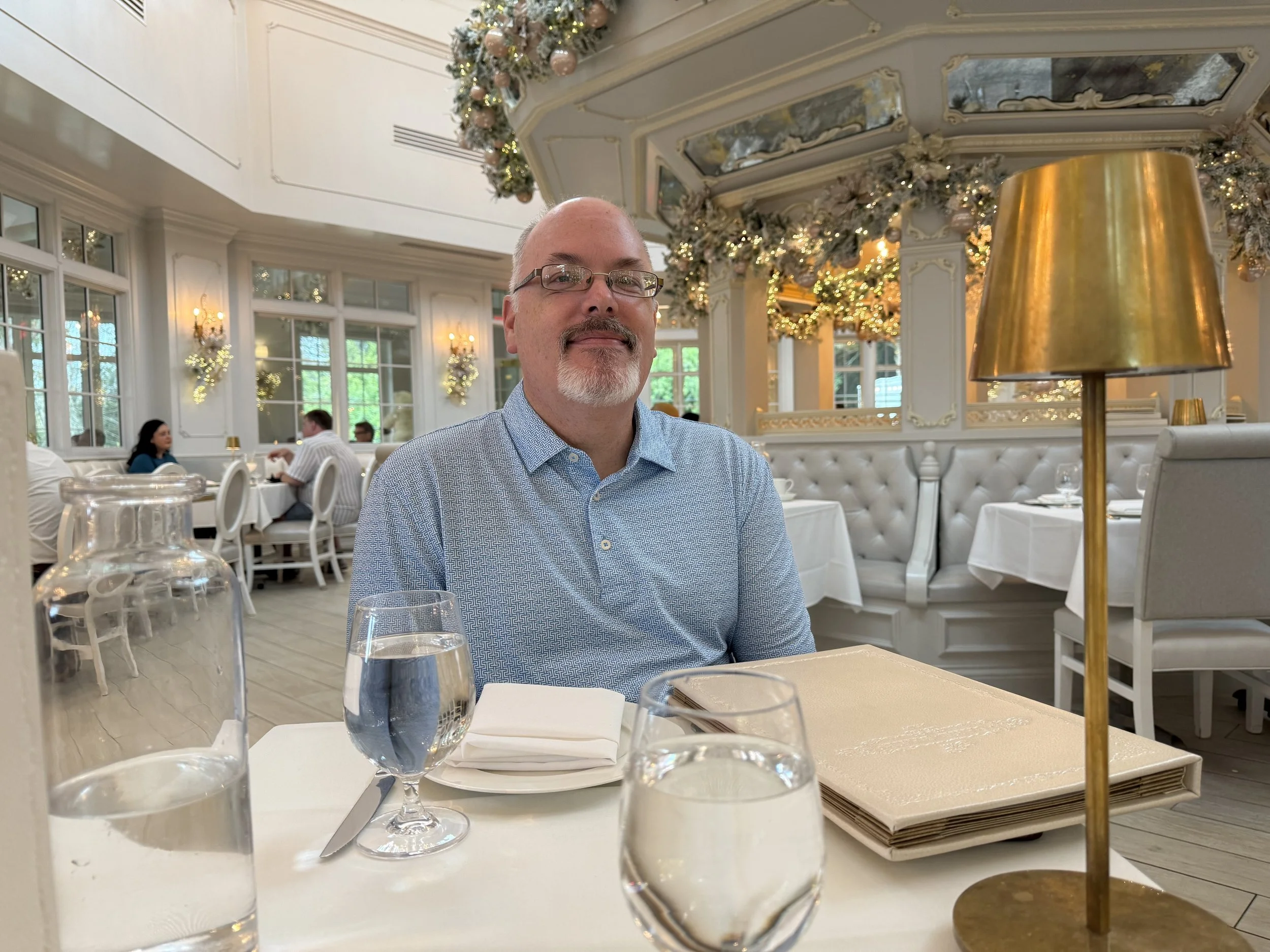 Man with glasses and a light blue shirt sitting at a dining table in a decorated elegant restaurant with festive holiday decor, including garlands and ornaments, seen through large windows.