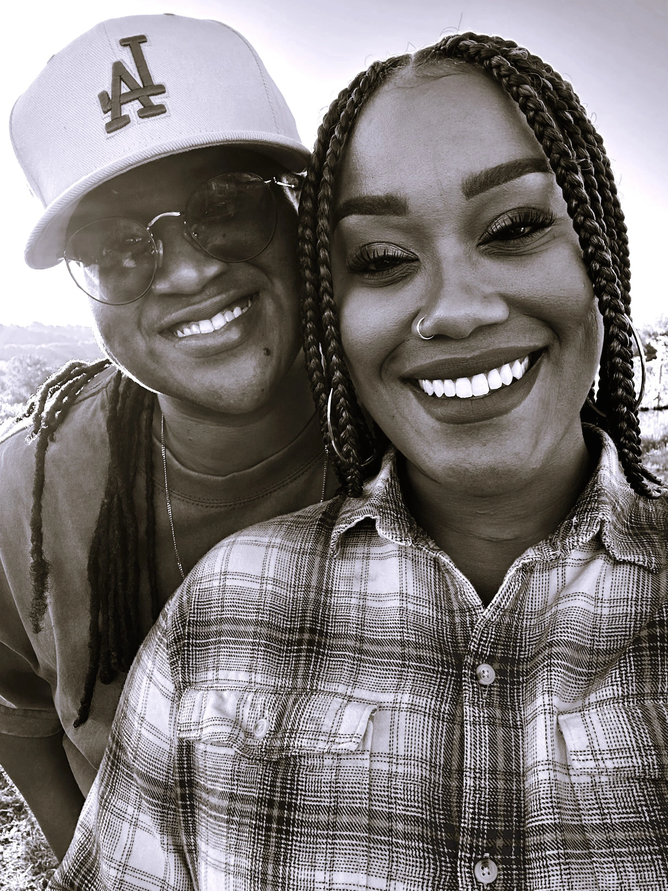 Two women taking a selfie together outdoors, smiling, with one wearing a Los Angeles Dodgers baseball cap and sunglasses, and the other wearing a plaid shirt and hoop earrings.