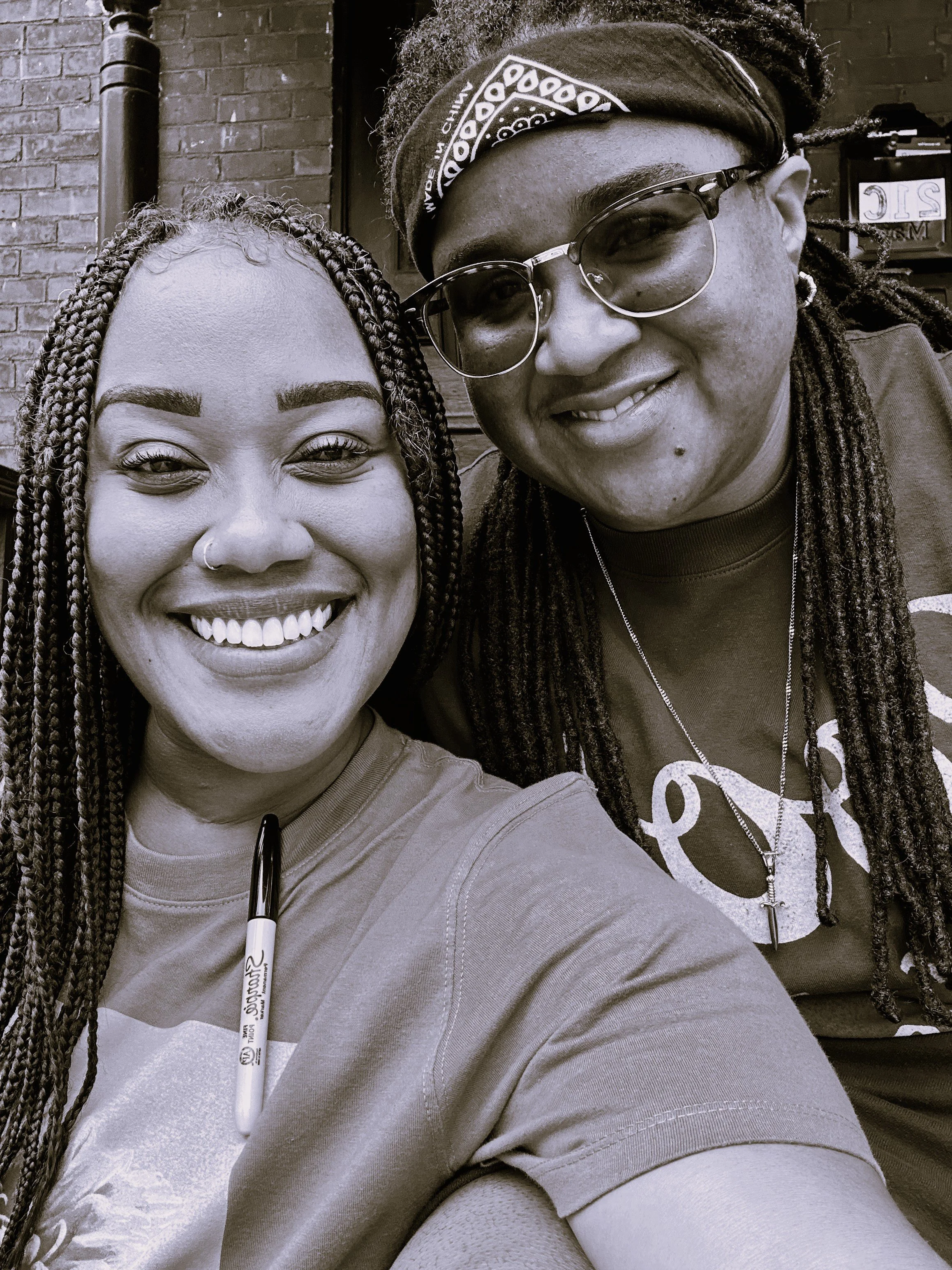 Close-up of two smiling people taking a selfie inside a building with a brick wall in the background. One person has braided hair and a nose ring, the other is wearing glasses and a bandana.