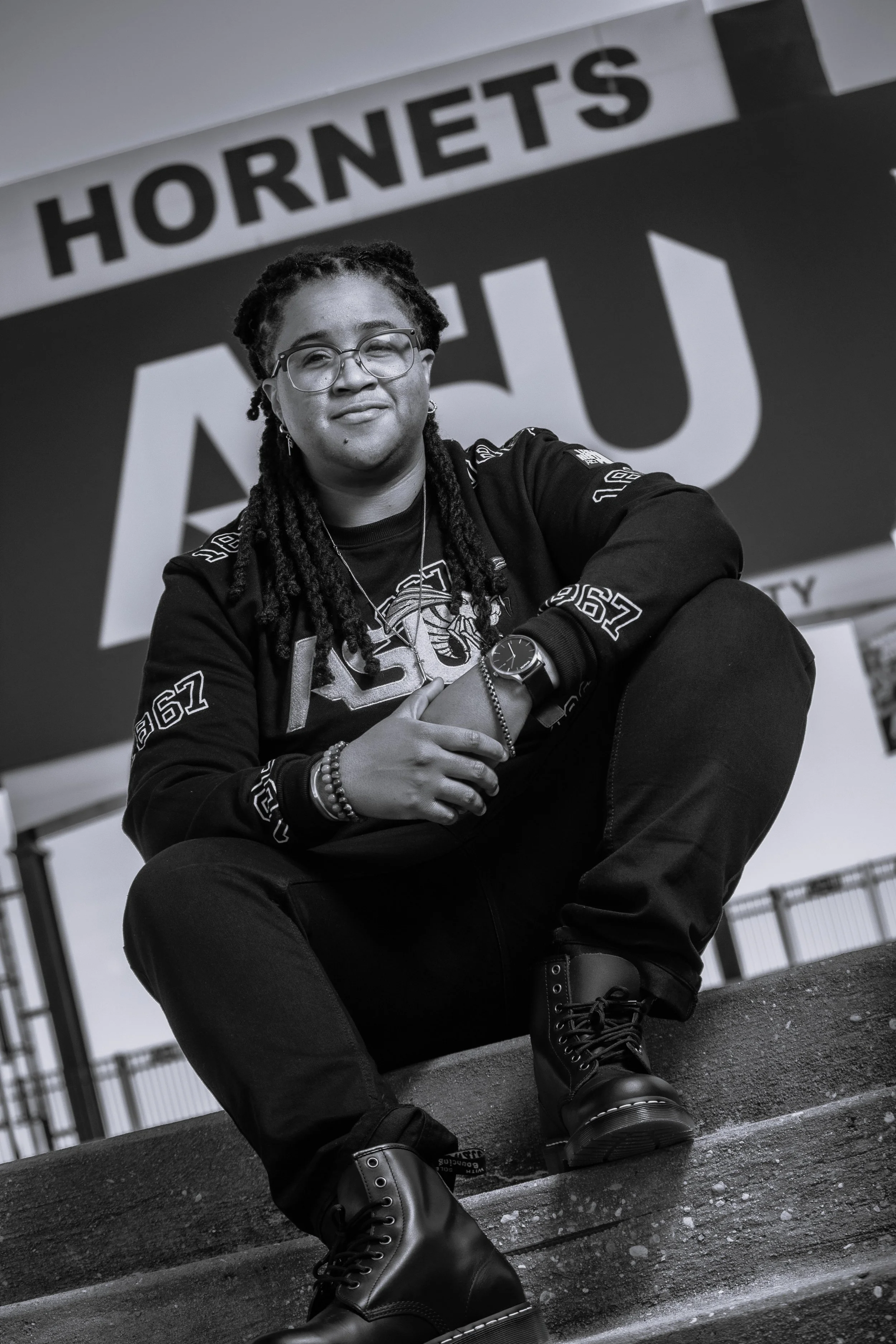 A person with glasses and dreadlocks sitting on stairs in front of a sign that reads 'HORNETS.'
