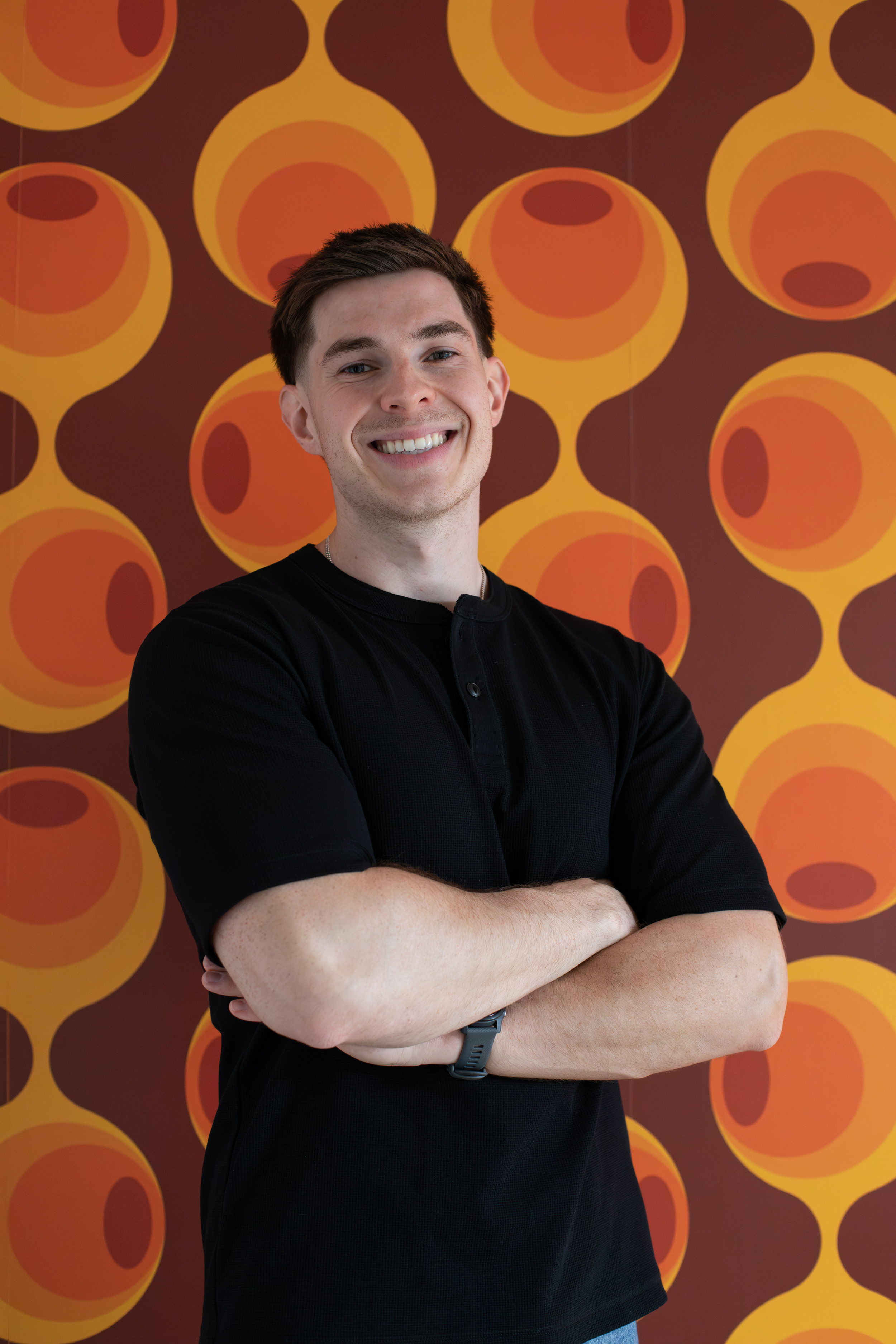 Un jeune homme souriant avec les bras croisés, portant une montre connectée, se tient devant un mur décoré de grands cercles oranges et jaunes.