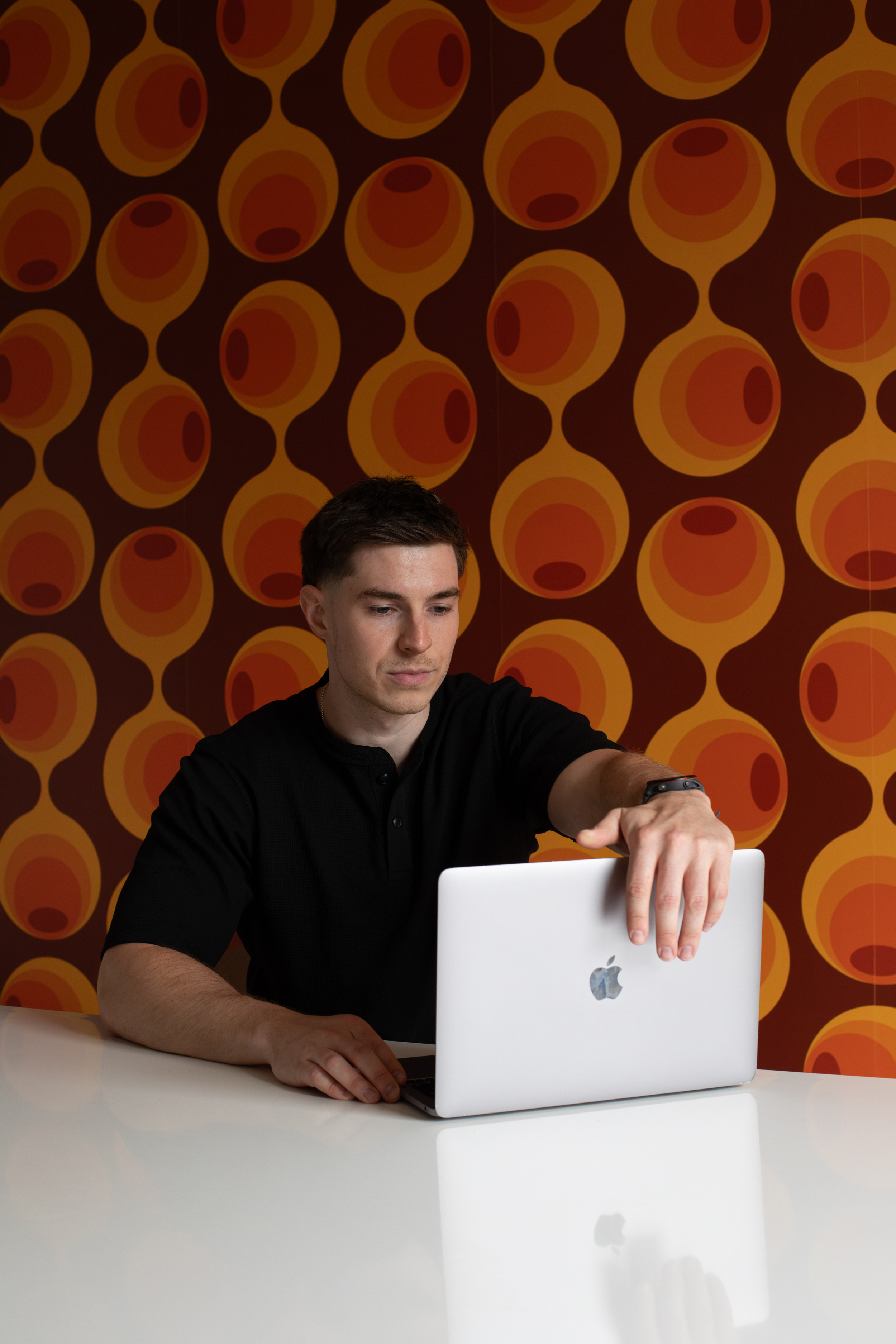 Un jeune homme assis à une table blanche utilisant un ordinateur portable blanc avec un fond de mur orné d'un motif rétro de cercles orange et marron.