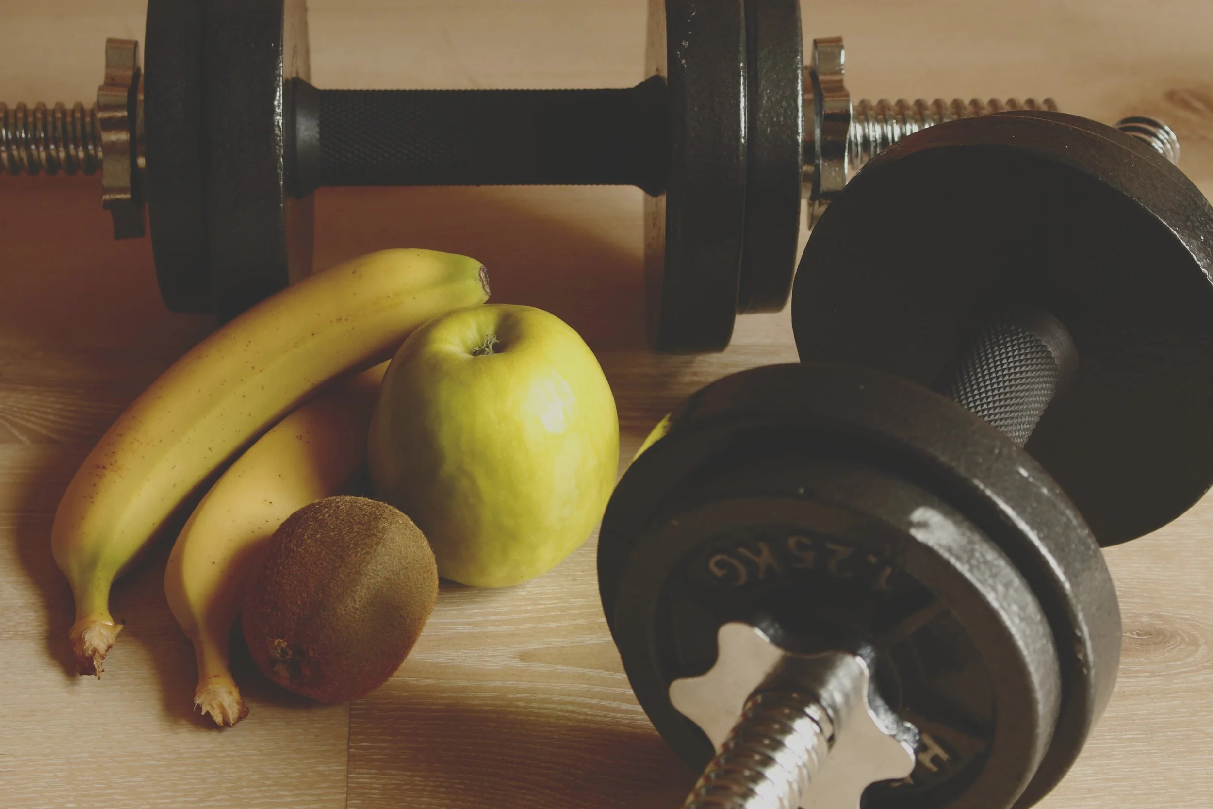 Des haltères, des bananes, une pomme, deux kiwis et un haltère de musculation sur une surface en bois.