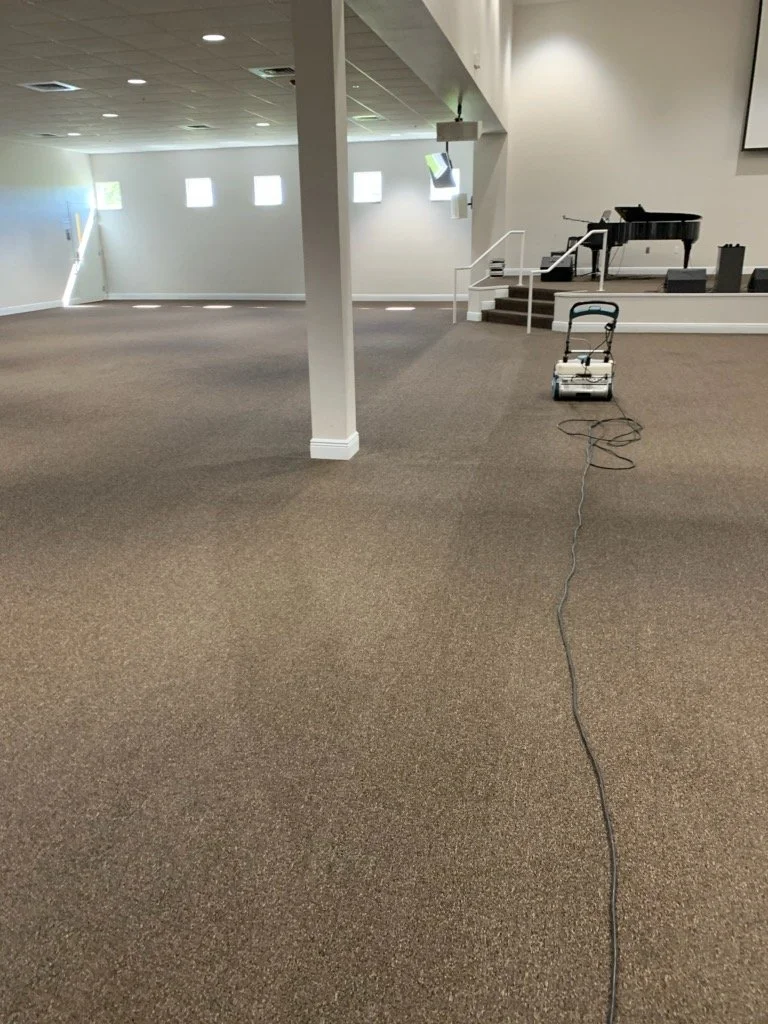 Empty room with carpet flooring, a small stage with a piano, a chair, and a cleaning machine.