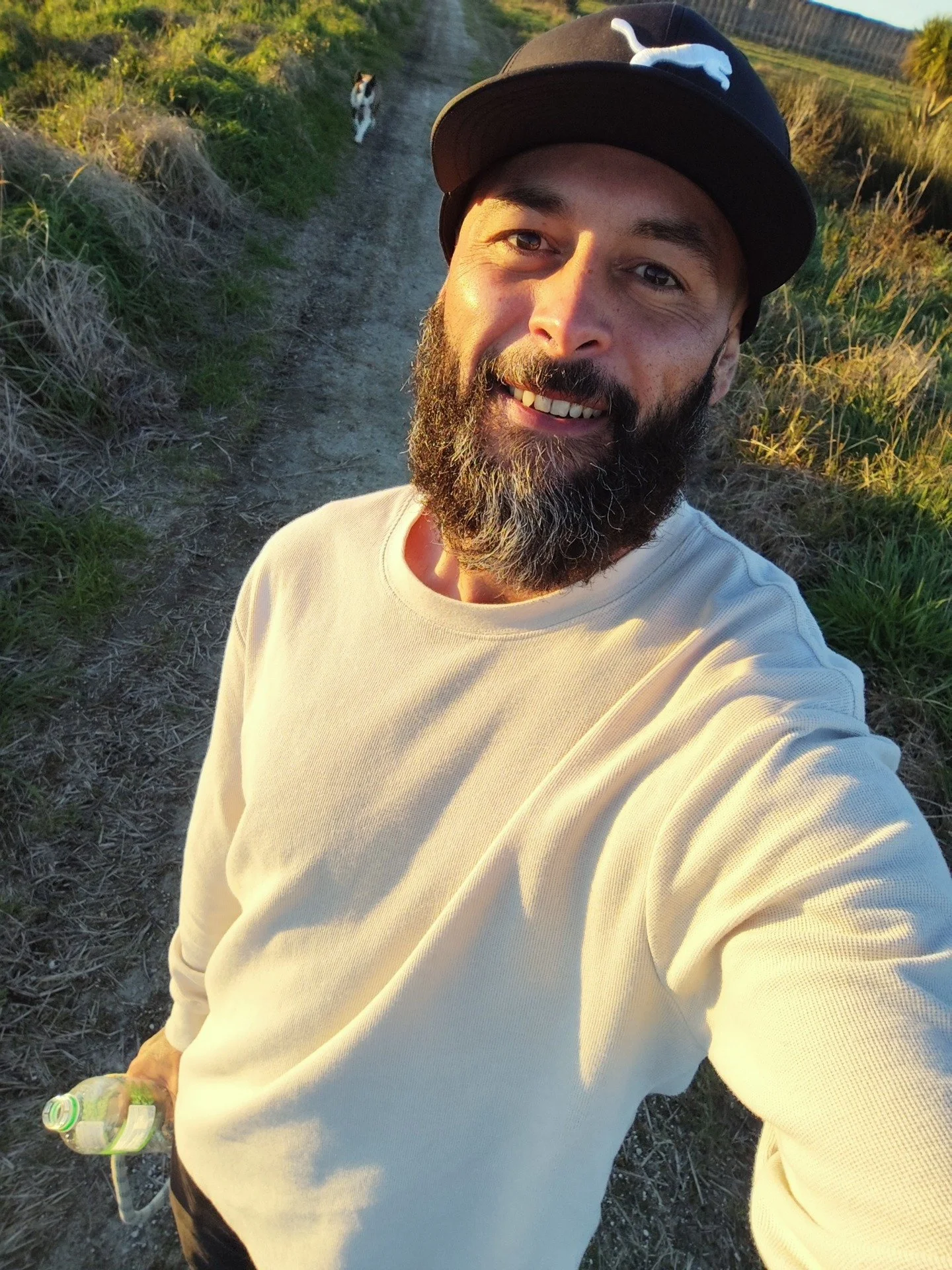 Bearded man in a black cap and beige long-sleeve shirt taking a selfie on a sunlit rural trail, holding a water bottle, with a bull terrier walking behind him beside a wire fence.