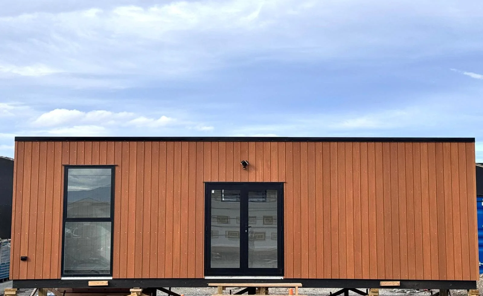A modern tiny house on stilts with brown vertical wood siding, black-framed glass door and window, situated outdoors on a partly cloudy day.