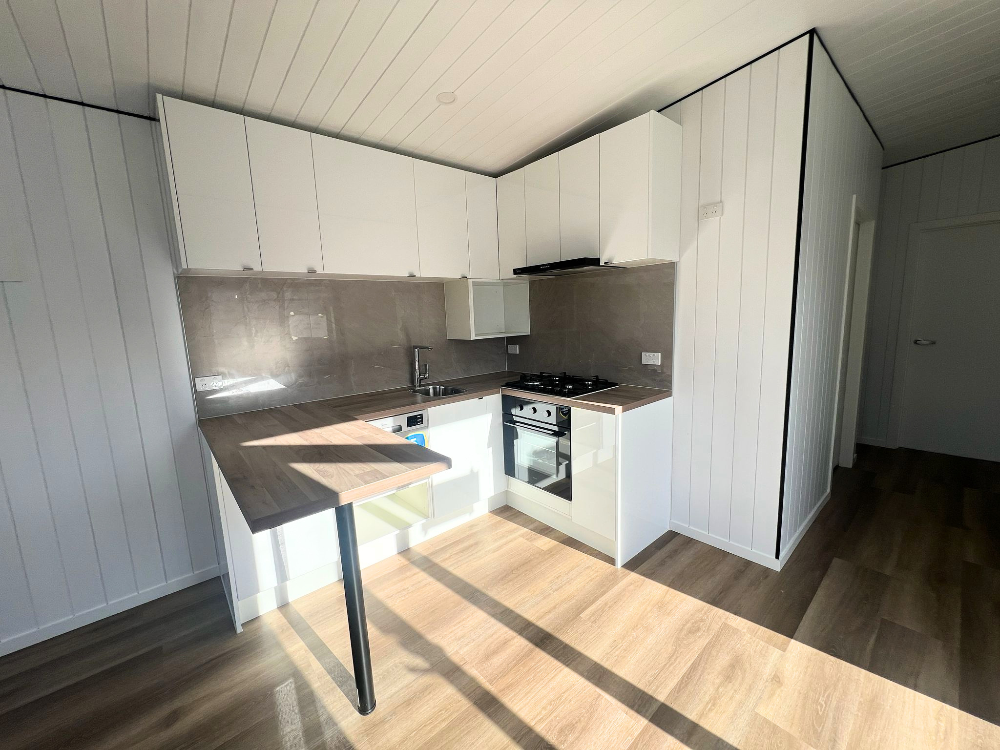 Modern kitchen with white cabinets, wooden countertop, built-in oven, gas stove, stainless steel sink, and beige tiled backsplash. White paneled walls and wooden flooring.