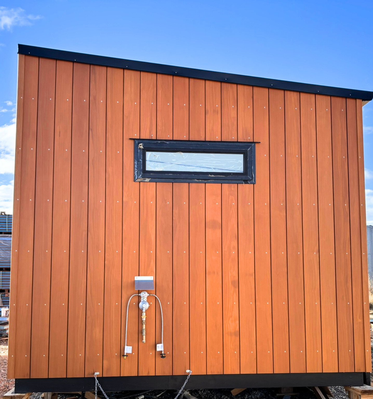 A modern tiny house with vertical wooden siding, a small horizontal window, and plumbing fixtures visible on the exterior against a clear blue sky.