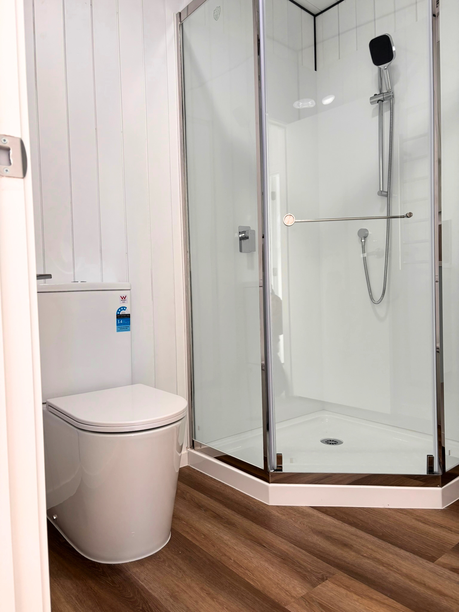 A modern bathroom featuring a white toilet and a corner shower with glass doors, chrome fixtures, and wood-look flooring.