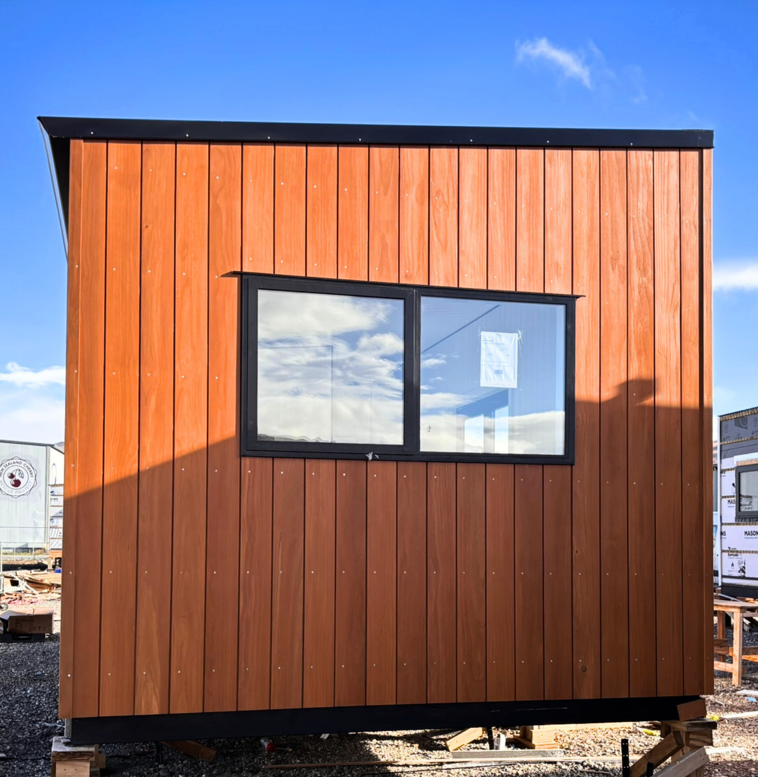 A small modern wooden building under construction with vertical wooden siding and a large rectangular window, set against a clear blue sky.