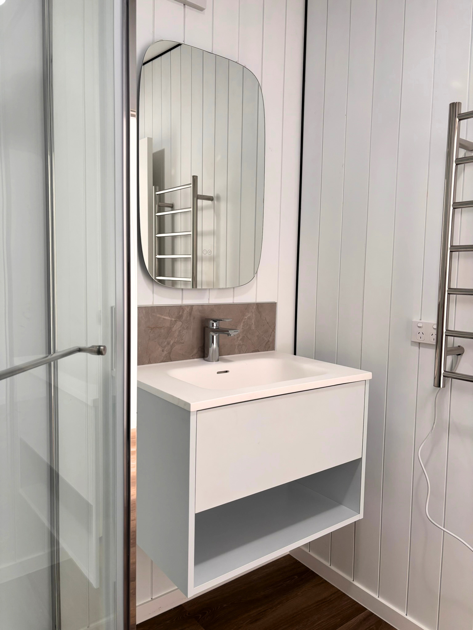 A bathroom vanity with a white cabinet, a beige countertop, a silver faucet, a wall-mounted mirror, and a partial view of a shower enclosure with a metal ladder-style towel rack. Wall panels are white vertically aligned, and the floor is dark wood.