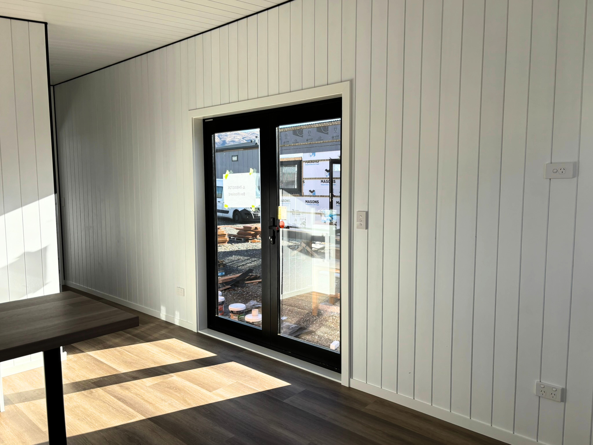 Interior view of a room with white paneled walls, a black-framed glass door, and a window showing construction outside. There's a wooden floor and a dark brown table.