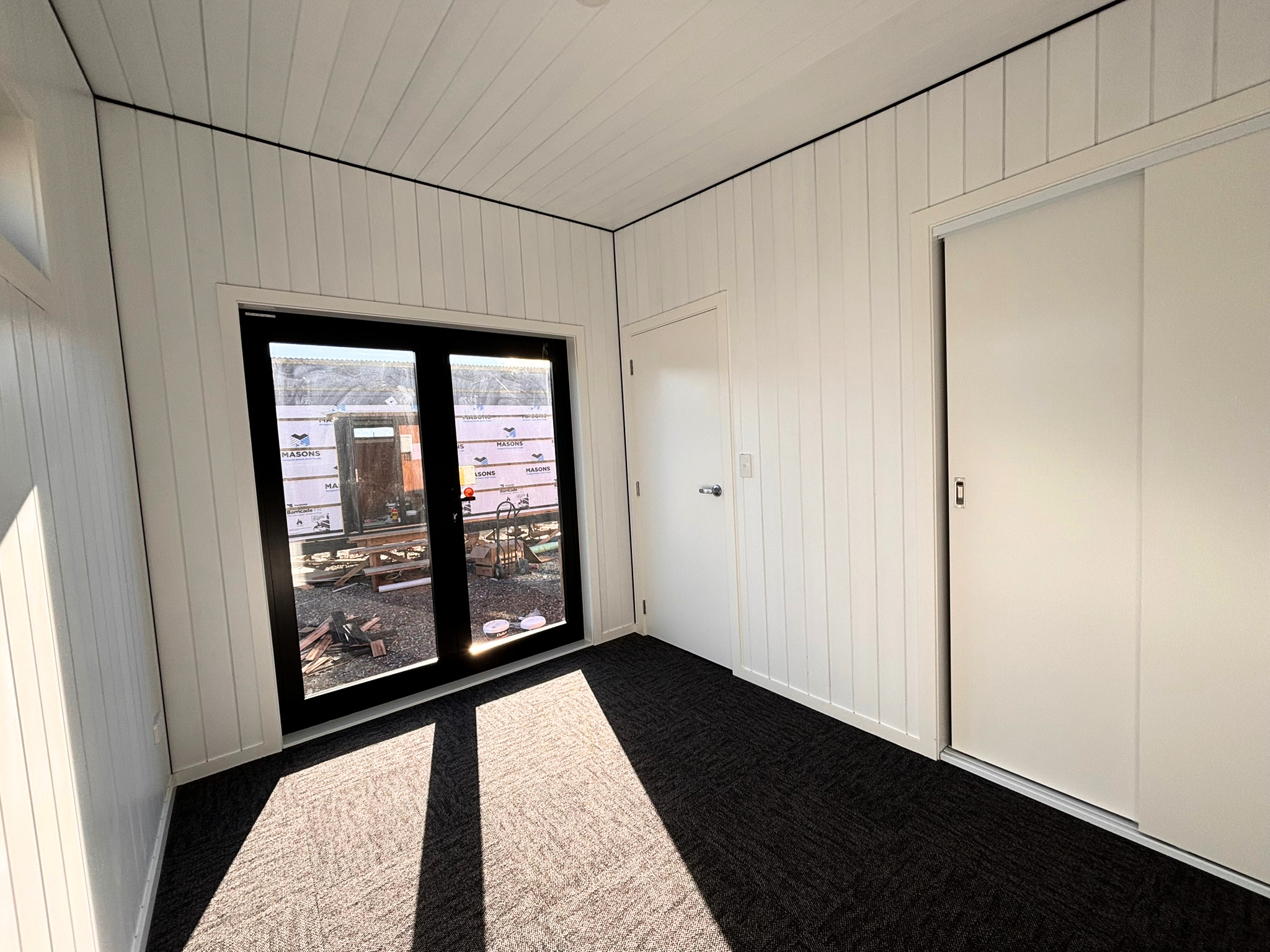 Interior of a room under construction with white beadboard walls, a black-framed window, and sliding closet doors. The floor features a black and beige patterned carpet, with construction materials visible outside the window.