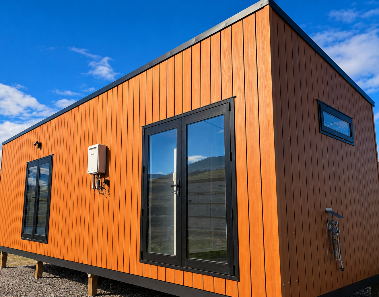 A modern tiny house with wooden exterior siding, black-framed glass doors, small rectangular windows, and outdoor utility fixtures under a blue sky with clouds.
