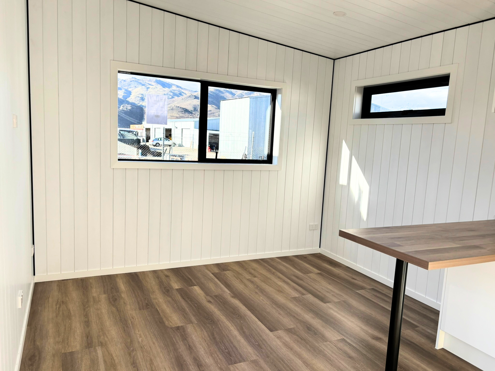 Interior of a room with white paneled walls, wood flooring, and large windows showing a mountain landscape outside. There is a wooden table with metal legs in the corner.