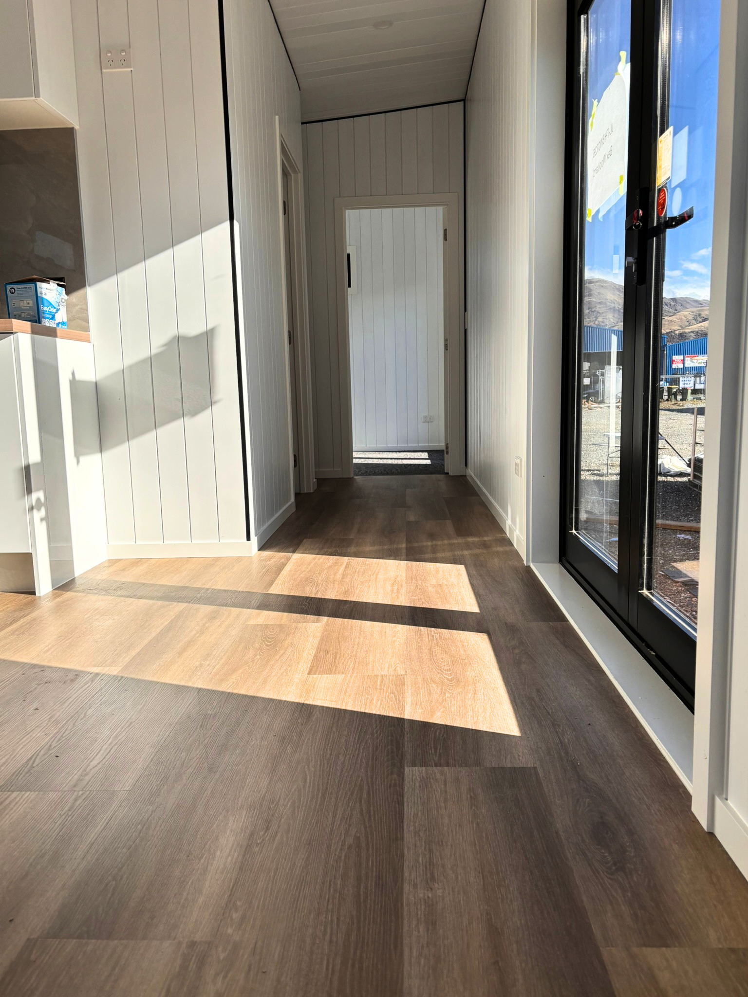 Interior of a modern building with wooden flooring, white paneled walls, and a large glass door leading outside, with mountains visible in the background.