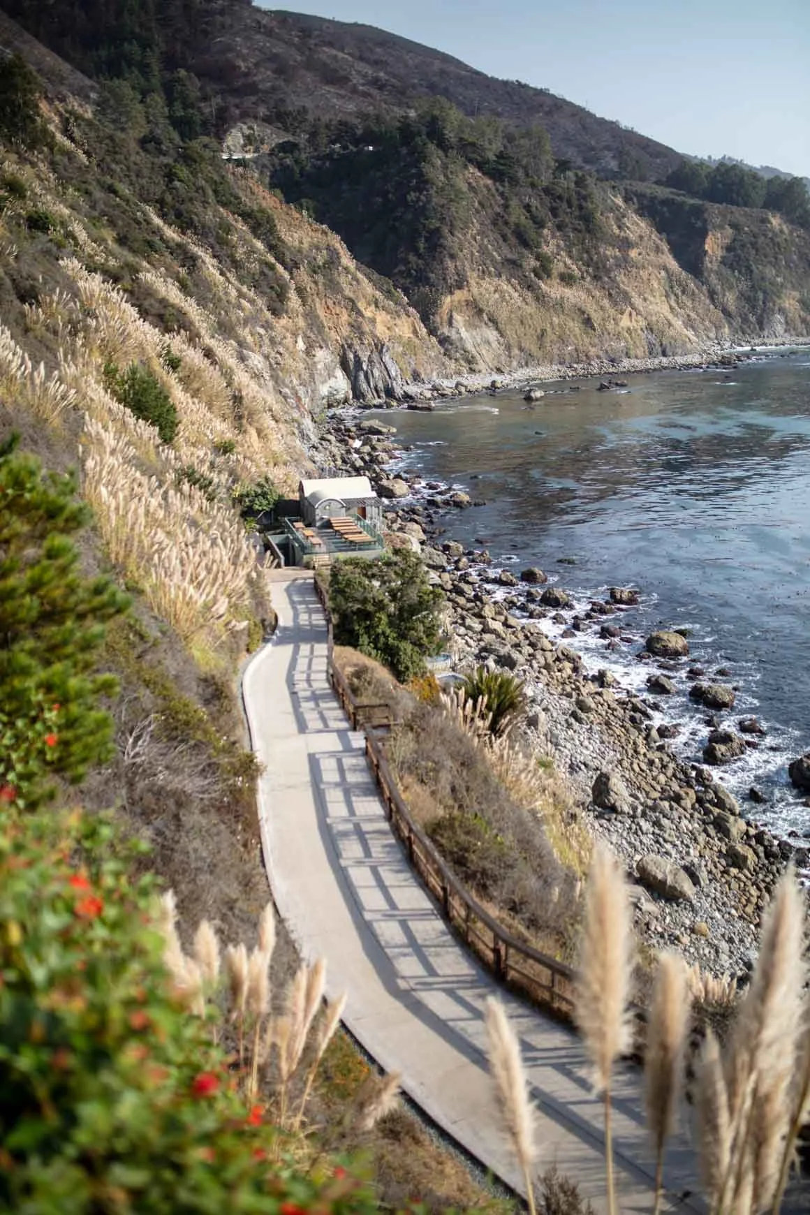 A winding concrete pathway along a rocky coastline with ocean waves, overlooking a hillside covered in grasses and shrubs.