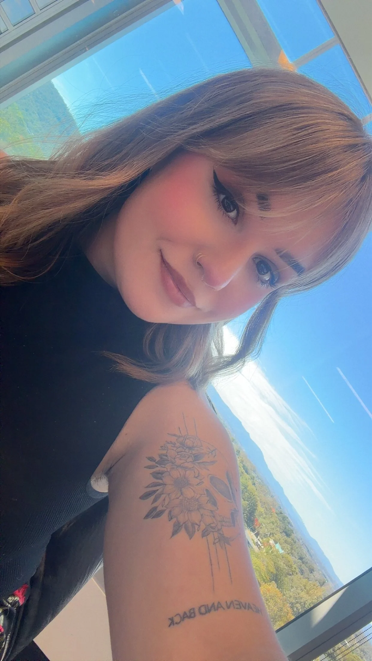 A young woman with brown hair and makeup taking a selfie with mountains and a blue sky in the background.
