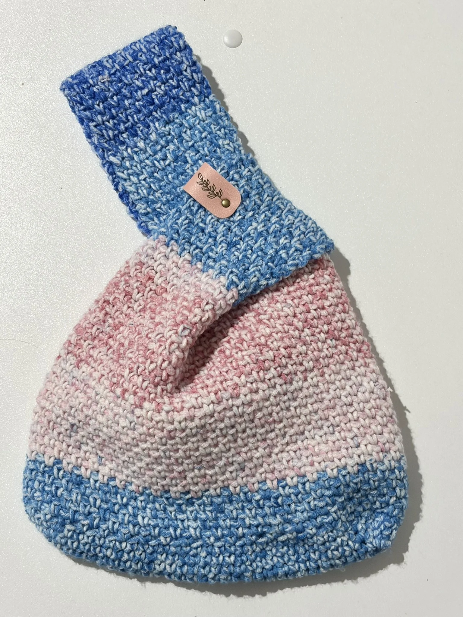 Close-up of a hand-knit pink and blue striped hat with a pink label featuring a black leaf design and a gold dot, placed on a white surface.