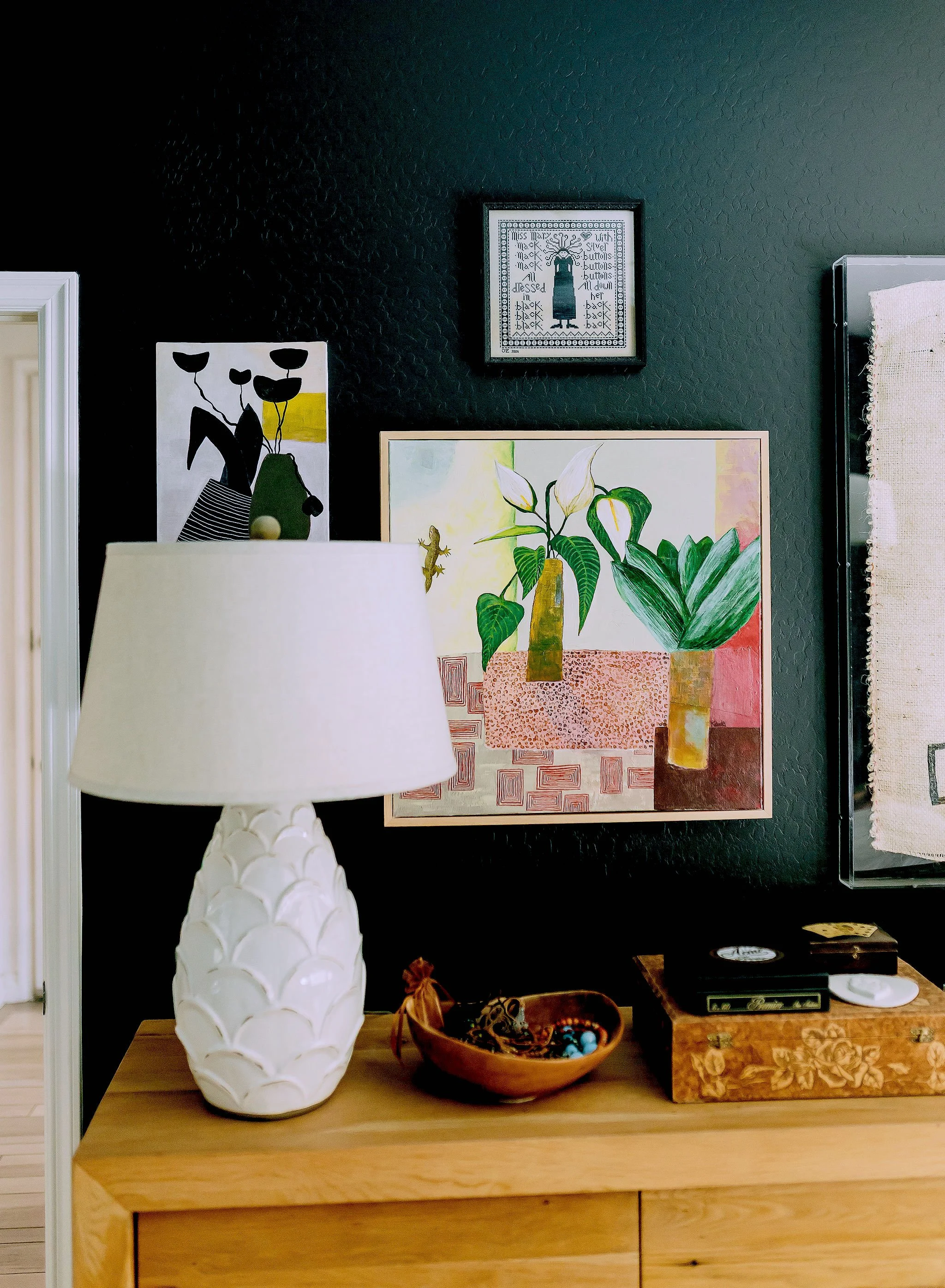 A wooden table with a white decorative lamp, a brown bowl filled with jewelry, and a wooden jewelry box on it. Behind the table, a dark-colored wall features framed artwork, including a framed cross-stitch pattern and a botanical illustration. Part o