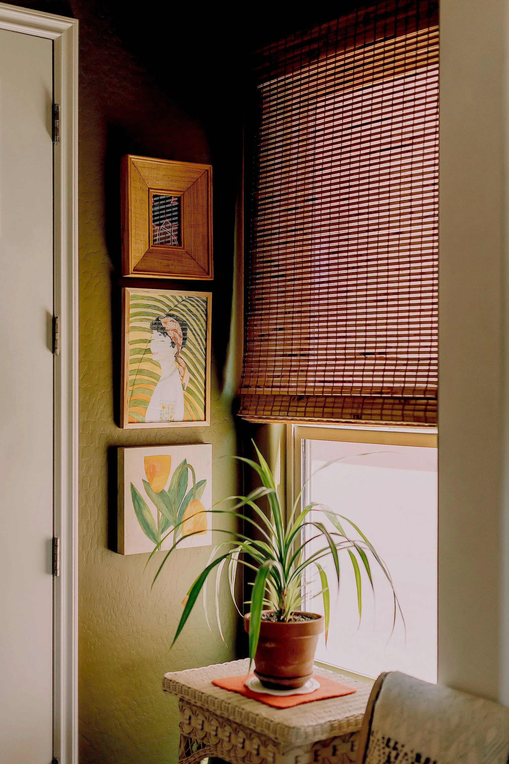 A room with a large potted plant on a wicker table, hanging art on a green textured wall, and closed bamboo blinds over a window.