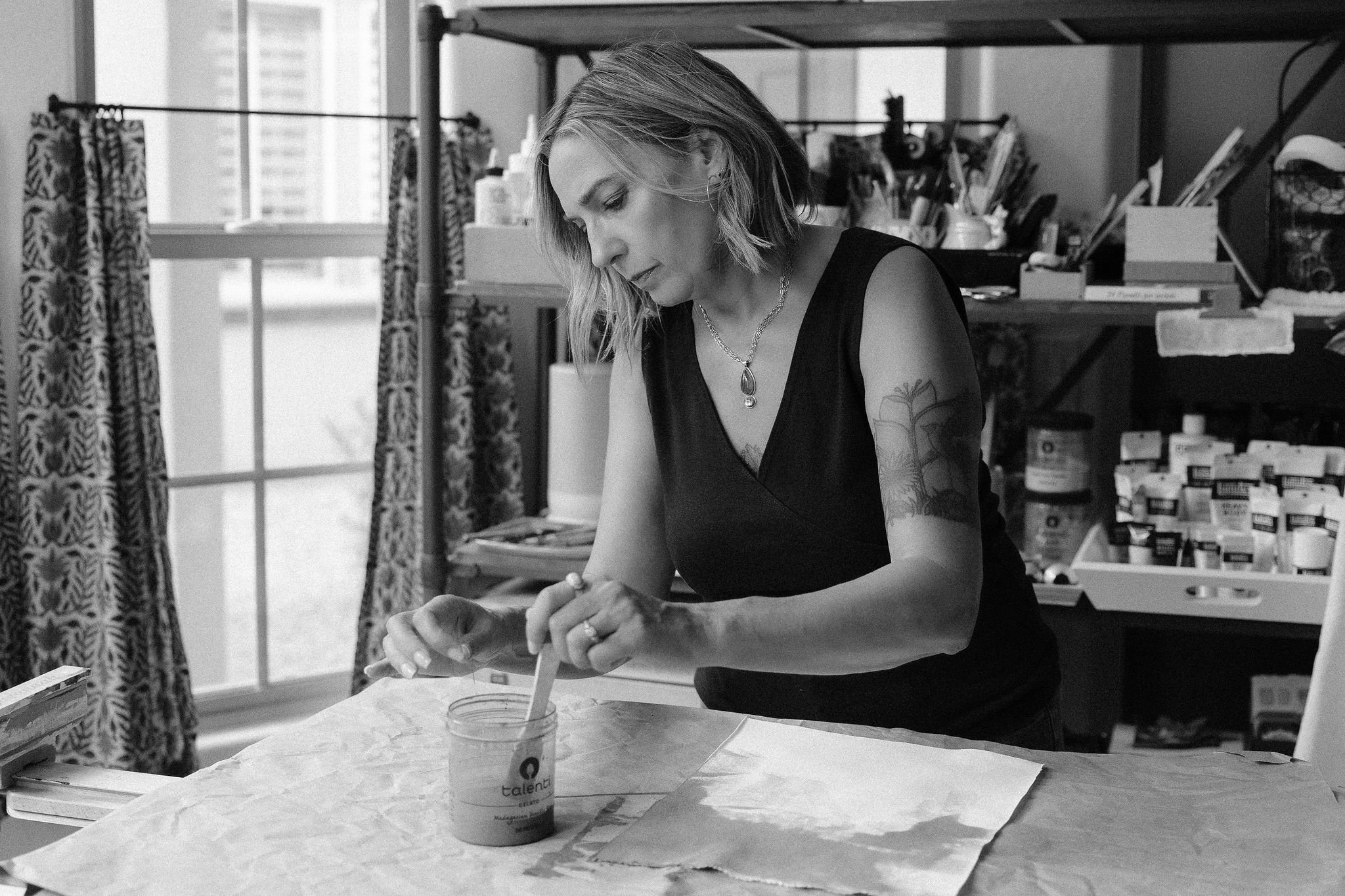 A woman with short hair and tattoos on her arms, wearing a black sleeveless top, working on a craft project at a table with art supplies in a room with large windows and patterned curtains.