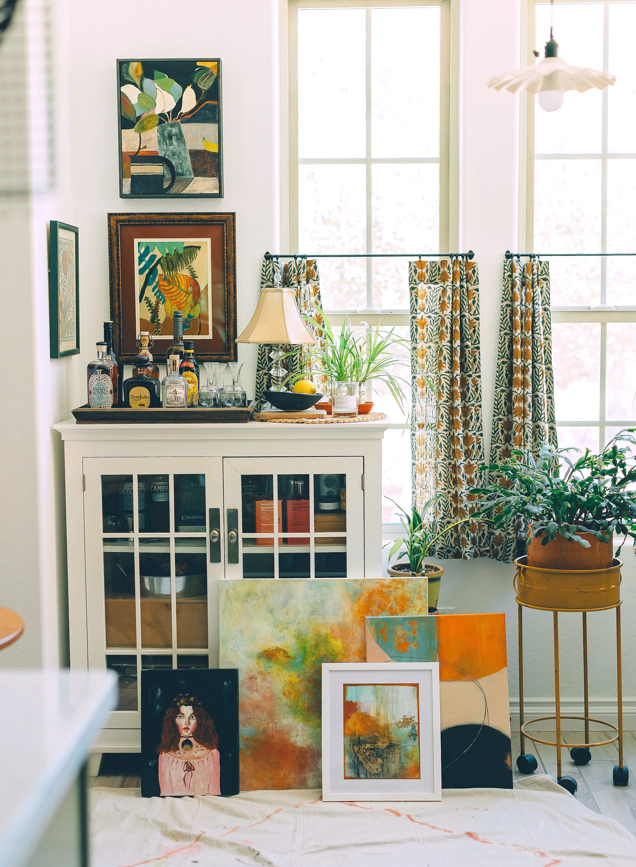A display of various colorful paintings arranged on a table and leaning against a white cabinet in front of a bright window with patterned curtains.