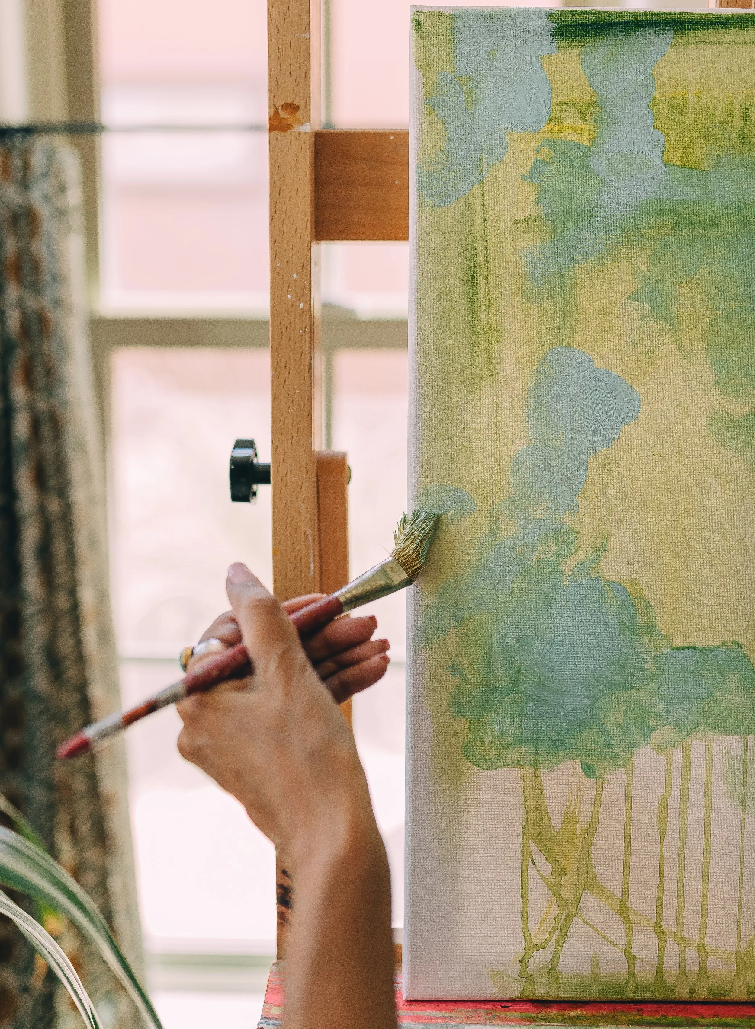 Person holding a paintbrush painting on a canvas with green and blue abstract colors, near a window with sunlight