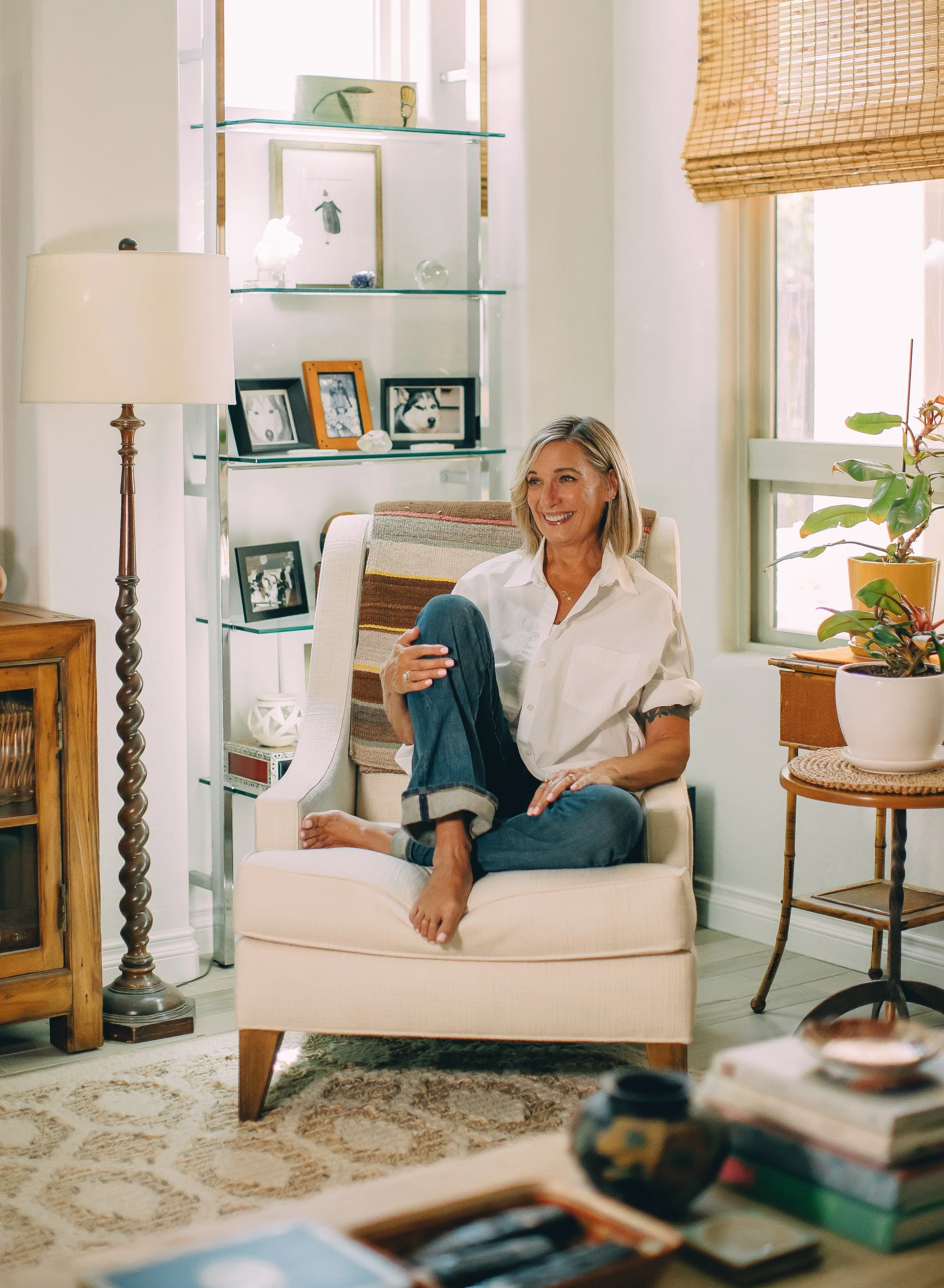 A woman sitting comfortably in an armchair in a well-lit living room, smiling. She has blonde hair, wears a white shirt, and dark jeans. A side table with a potted plant, a bookshelf with photographs, and a window with bamboo blinds are visible in the background.