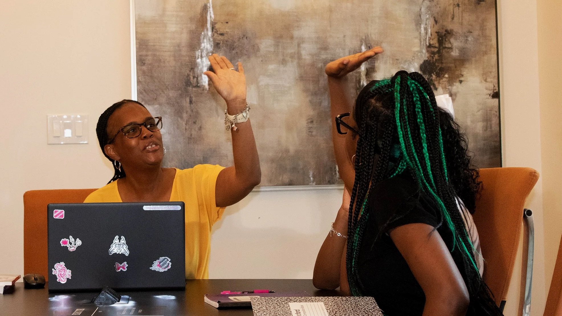 Two women sitting at a table, engaged in a conversation with one woman raising her hand. One woman has braids with green highlights and wears glasses, the other wears glasses and a yellow shirt. There are stickers on the laptop, notebooks, and a mouse on the table, with a large abstract painting in the background.