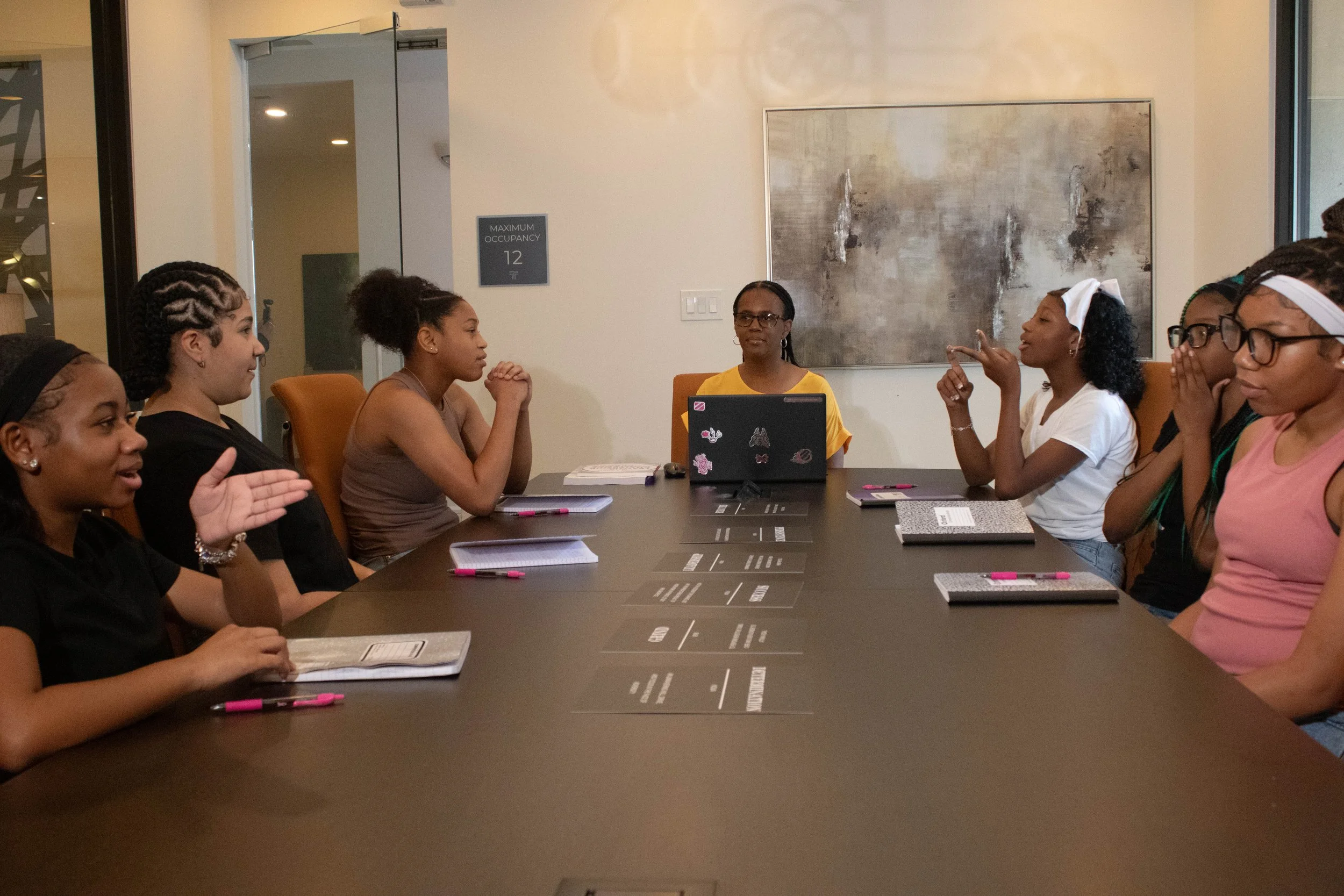 A group of women sitting around a conference table, engaging in discussion. There are notebooks, pens, and a laptop in front of them. The room has modern decor with abstract artwork on the wall.