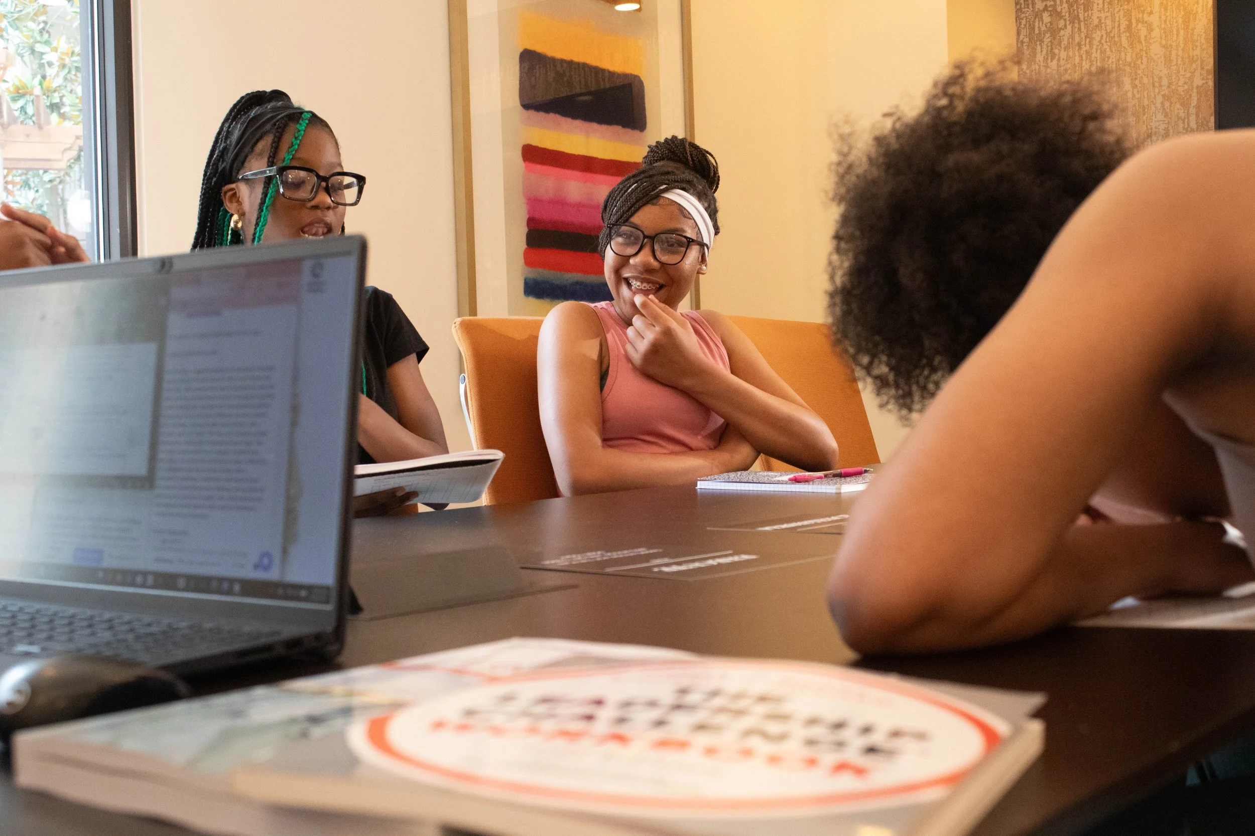 Four young women sitting around a table, smiling and engaging in conversation. One is wearing glasses with a head wrap, another has colorful braided hair and glasses, and the third has curly hair. There is a laptop, notebooks, and a cup on the table.