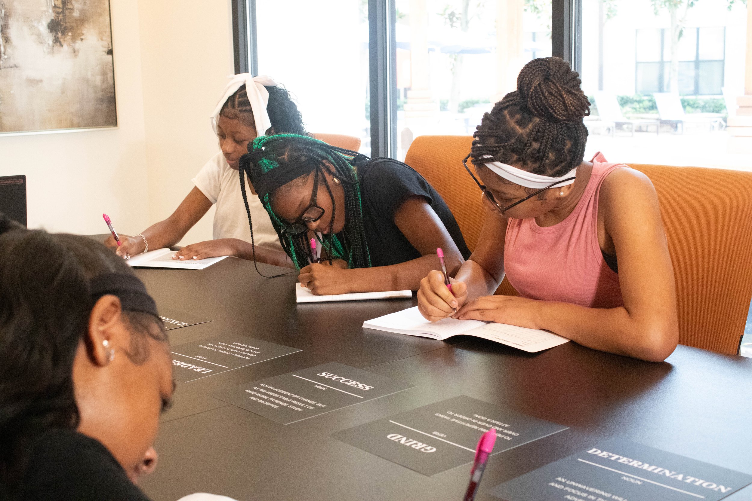 Three women with braided hair, wearing headbands, sitting at a table and writing in notebooks. They are in a well-lit room with a large window in the background.