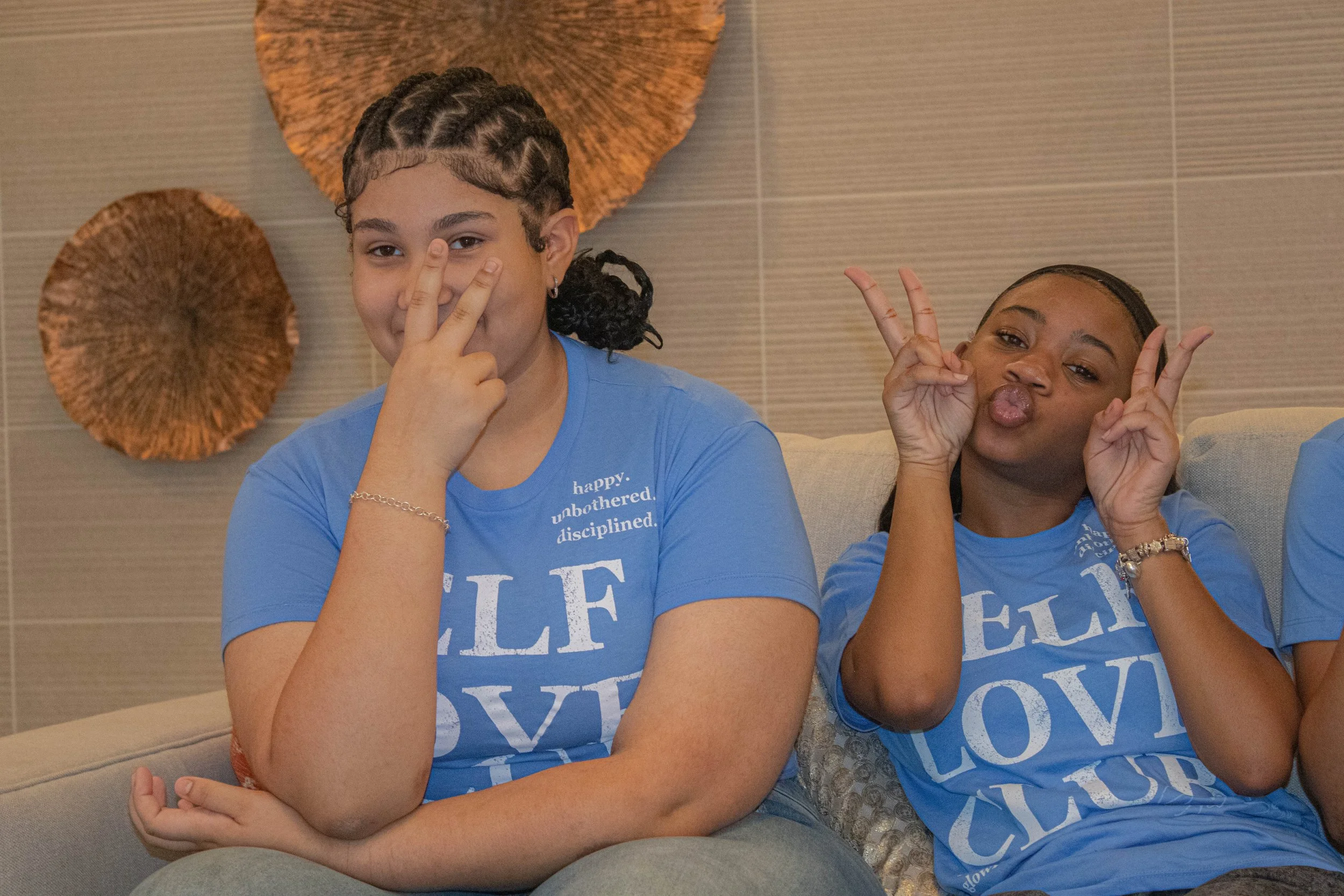 Two young women sitting on a beige couch, making playful gesture while smiling, in front of textured wall decor.