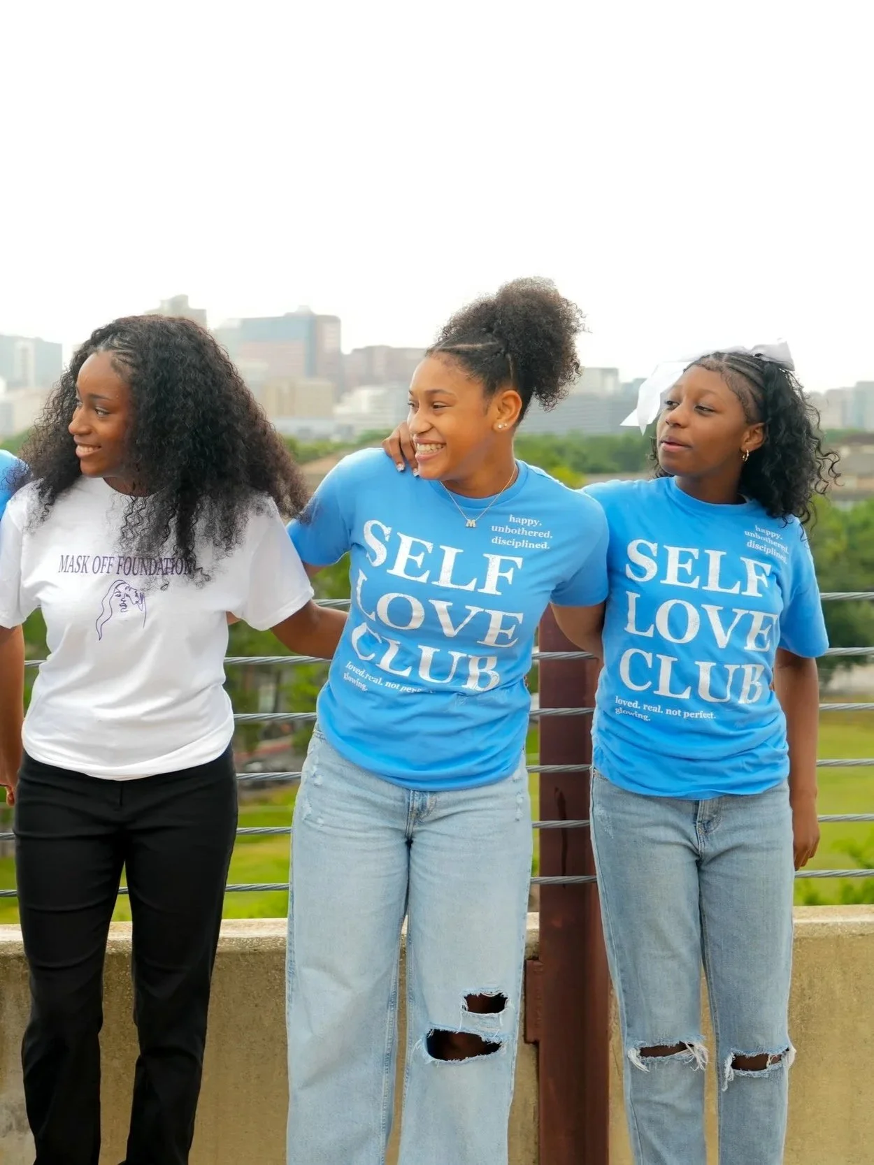 Three women standing outdoors against a city skyline, smiling and wearing blue and white T-shirts with phrases about self-love and happiness.